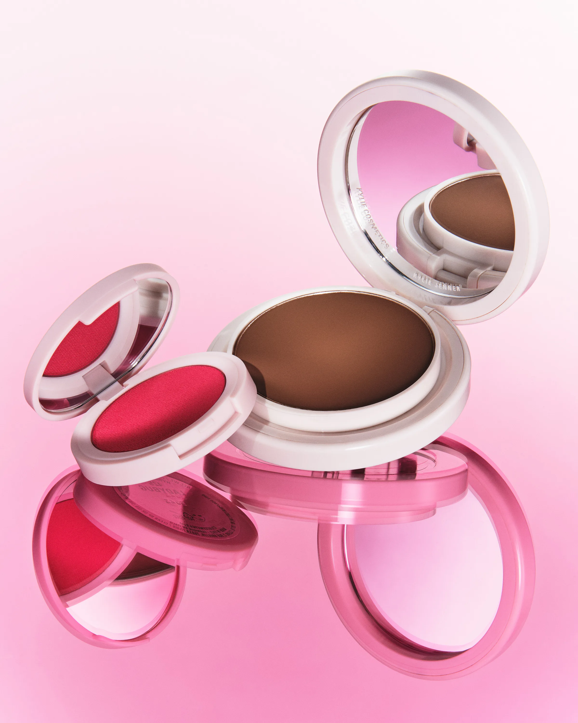 Open bronzer and red blush compacts with mirrors floating over a glossy pink reflective surface, for Kylie Cosmetics.