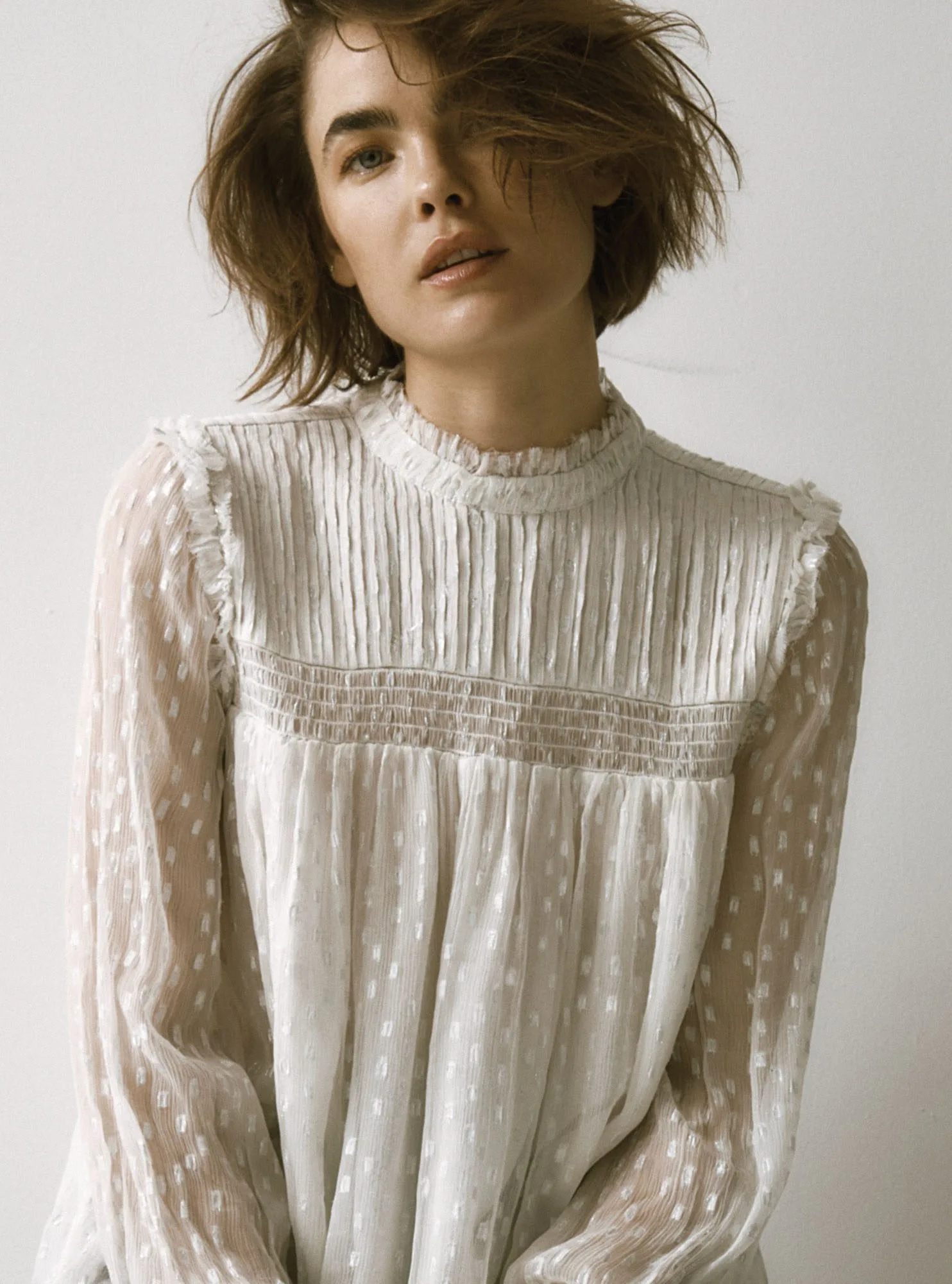 Model with tousled bob in a sheer white Swiss-dot blouse with pintucked ruffle yoke against a white wall, for FRAME.