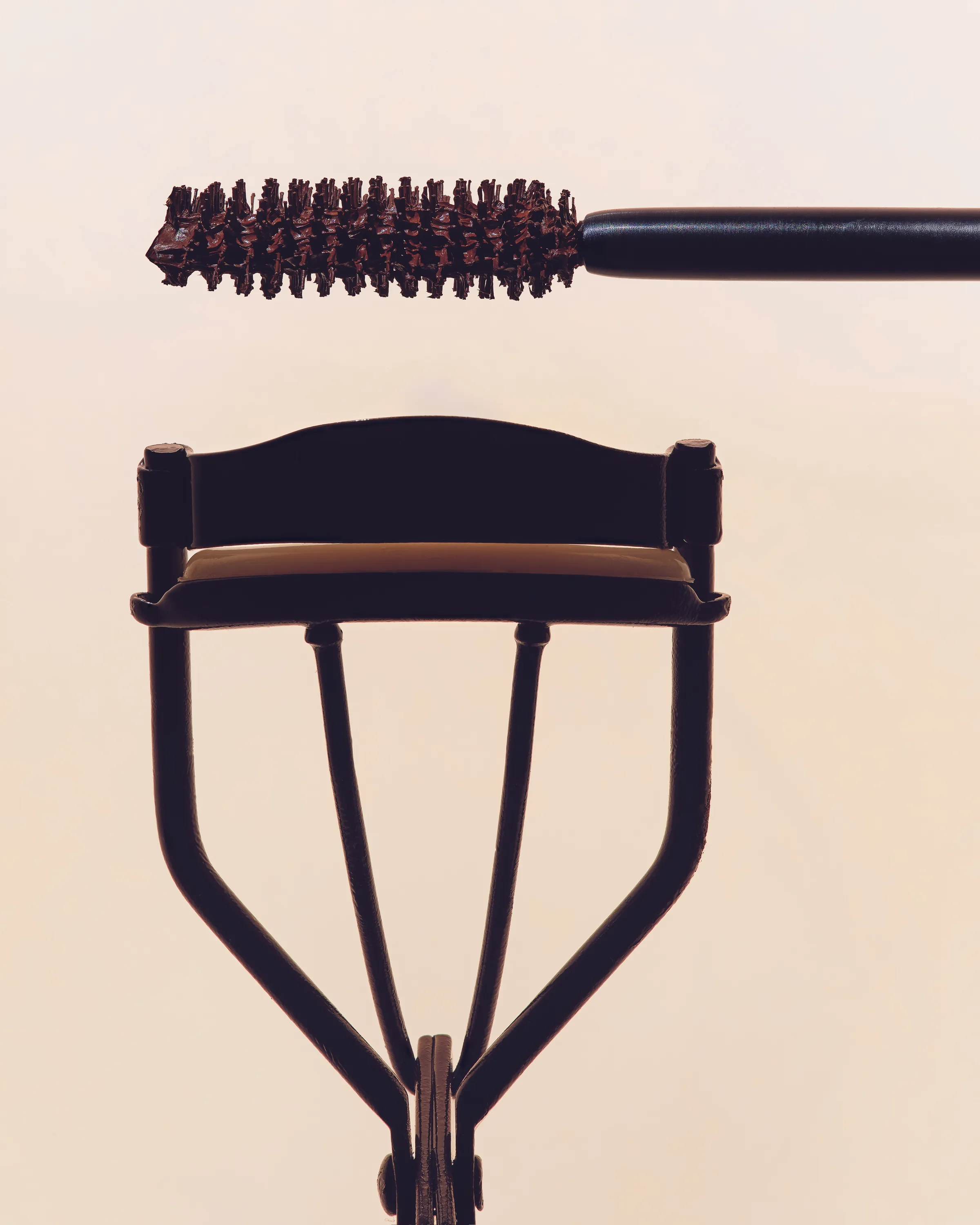 Brown mascara wand held above a black eyelash curler on a soft peach background, for Kylie Cosmetics.
