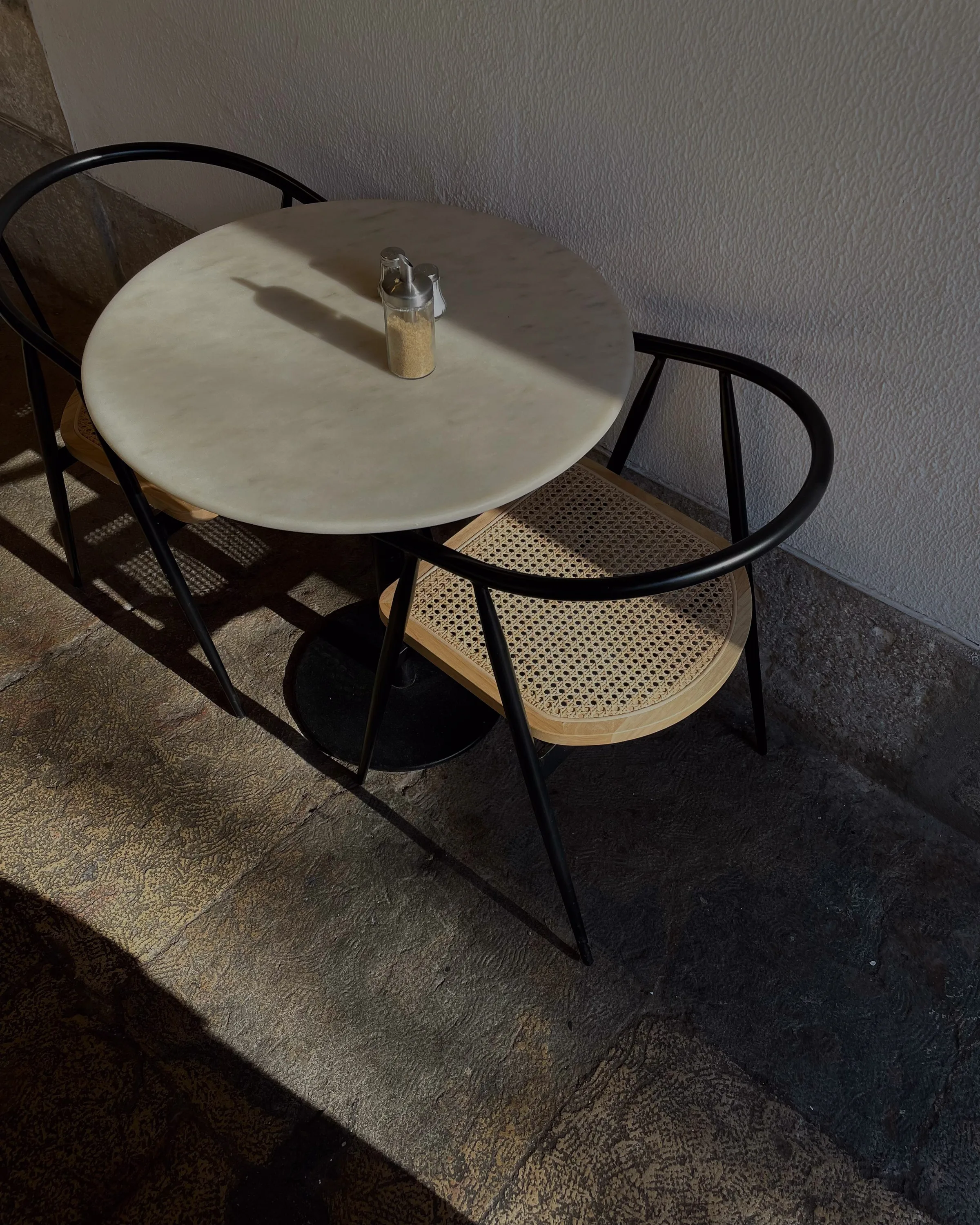 Round marble café table with two black cane-seat chairs against a white wall in low sunlight.