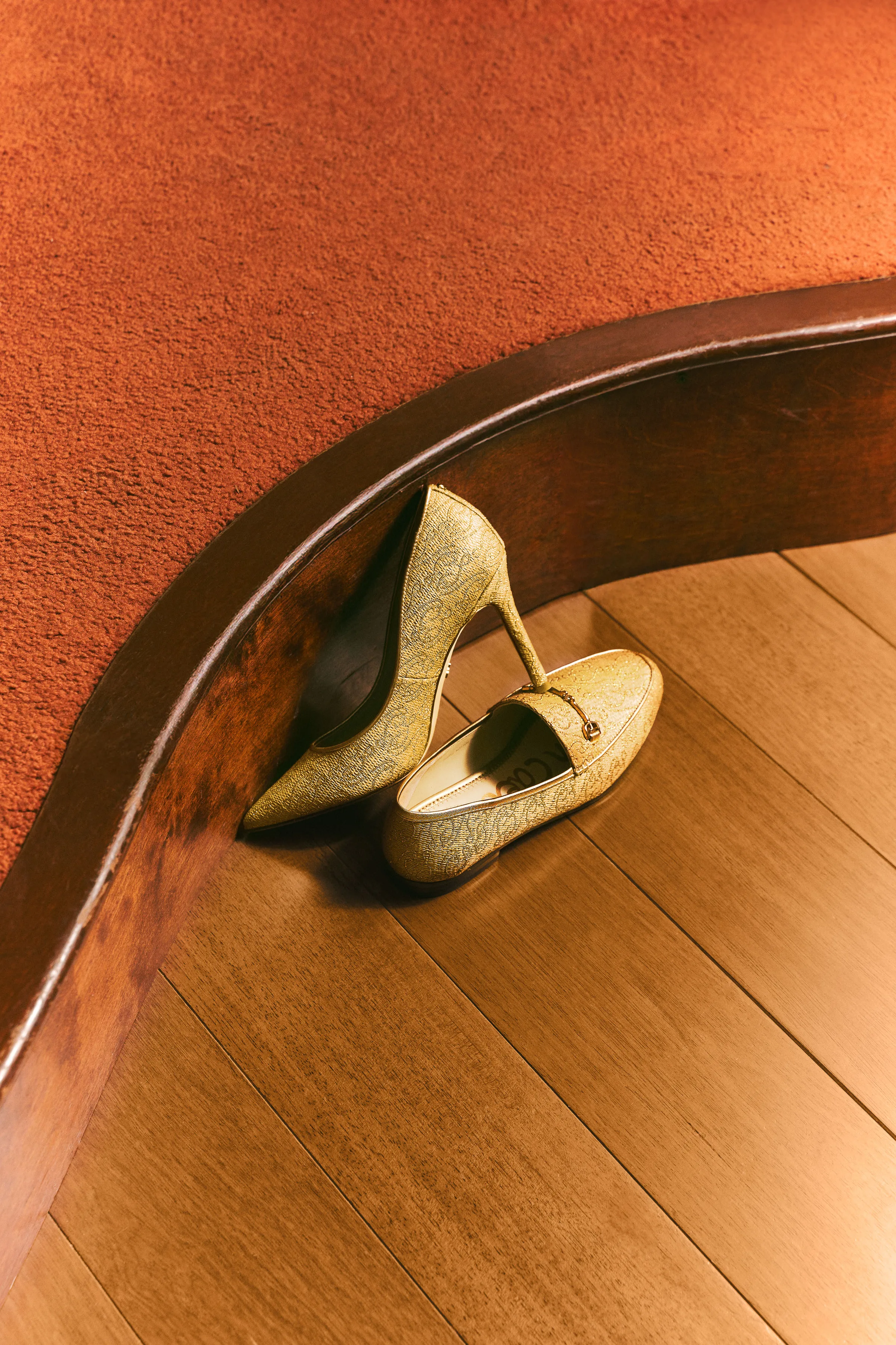 Gold textured stiletto pump leaning on a curved wood ledge beside a matching loafer on a wooden floor, for Sam Edelman.