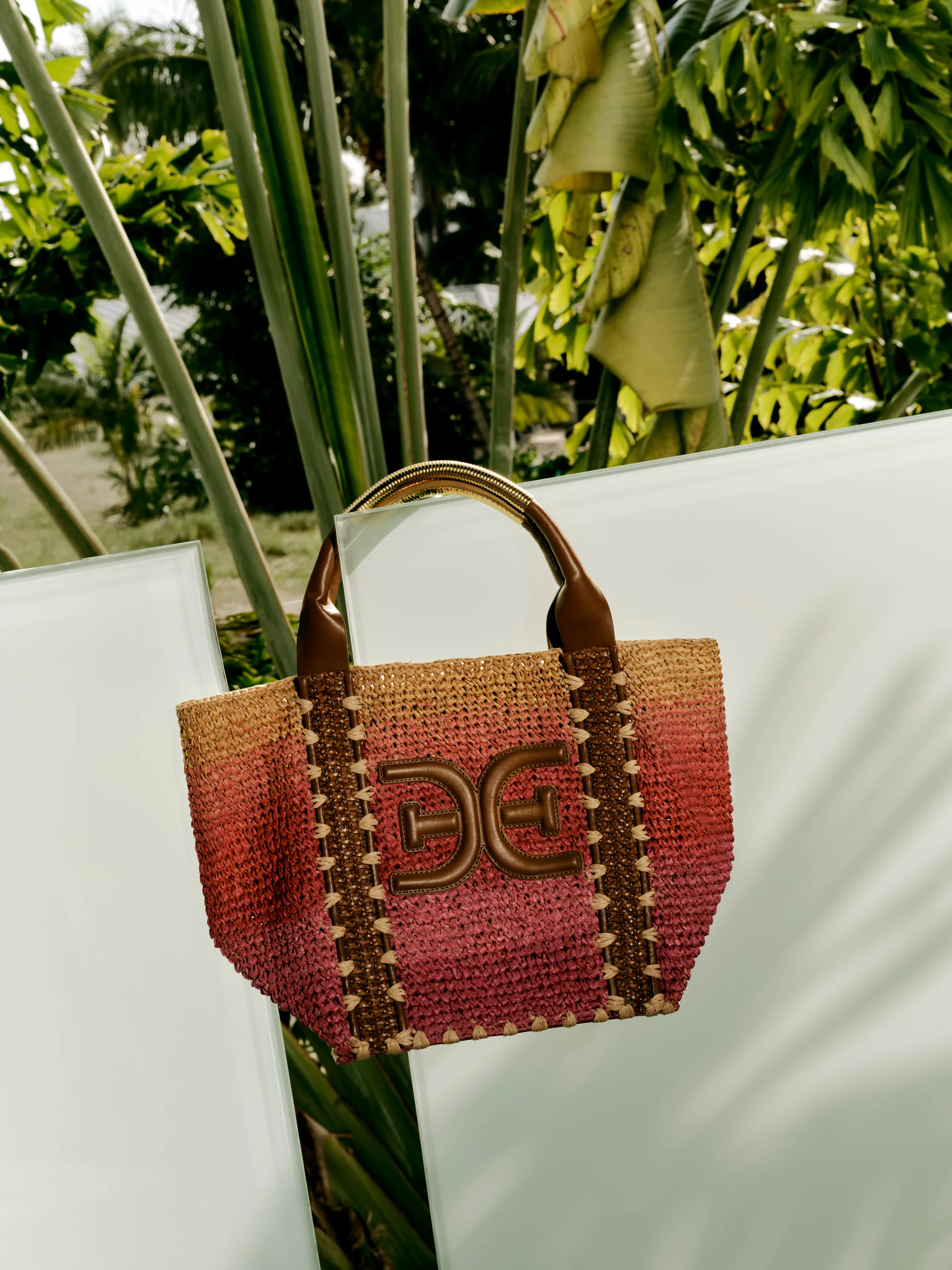 Pink and tan ombré raffia tote with leather handle and gold E logo, propped on glass panels among palms, for Sam Edelman.