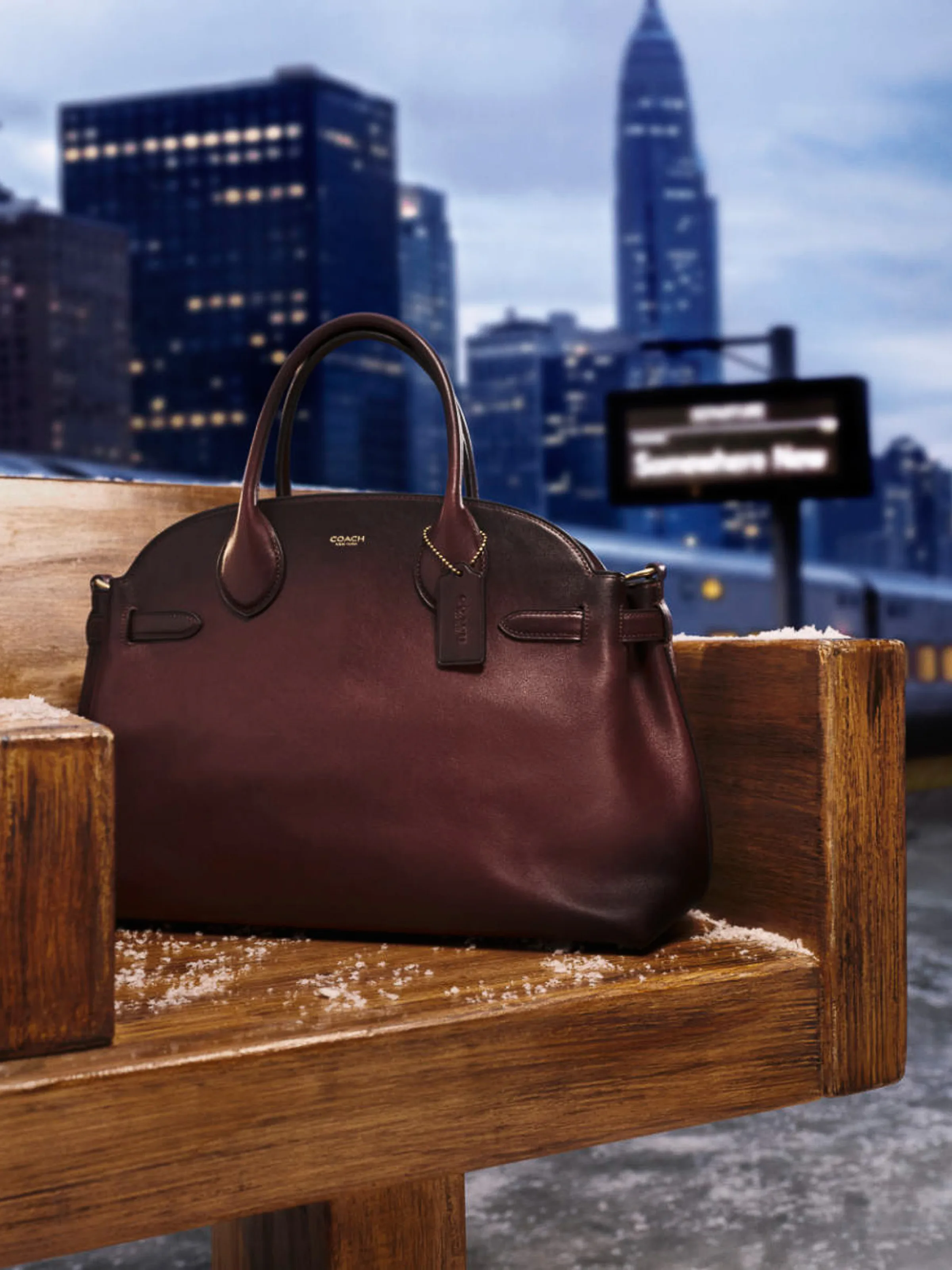 AI image of a burgundy leather handbag on a snowy wooden bench, New York skyline and train behind at dusk, for Coach.