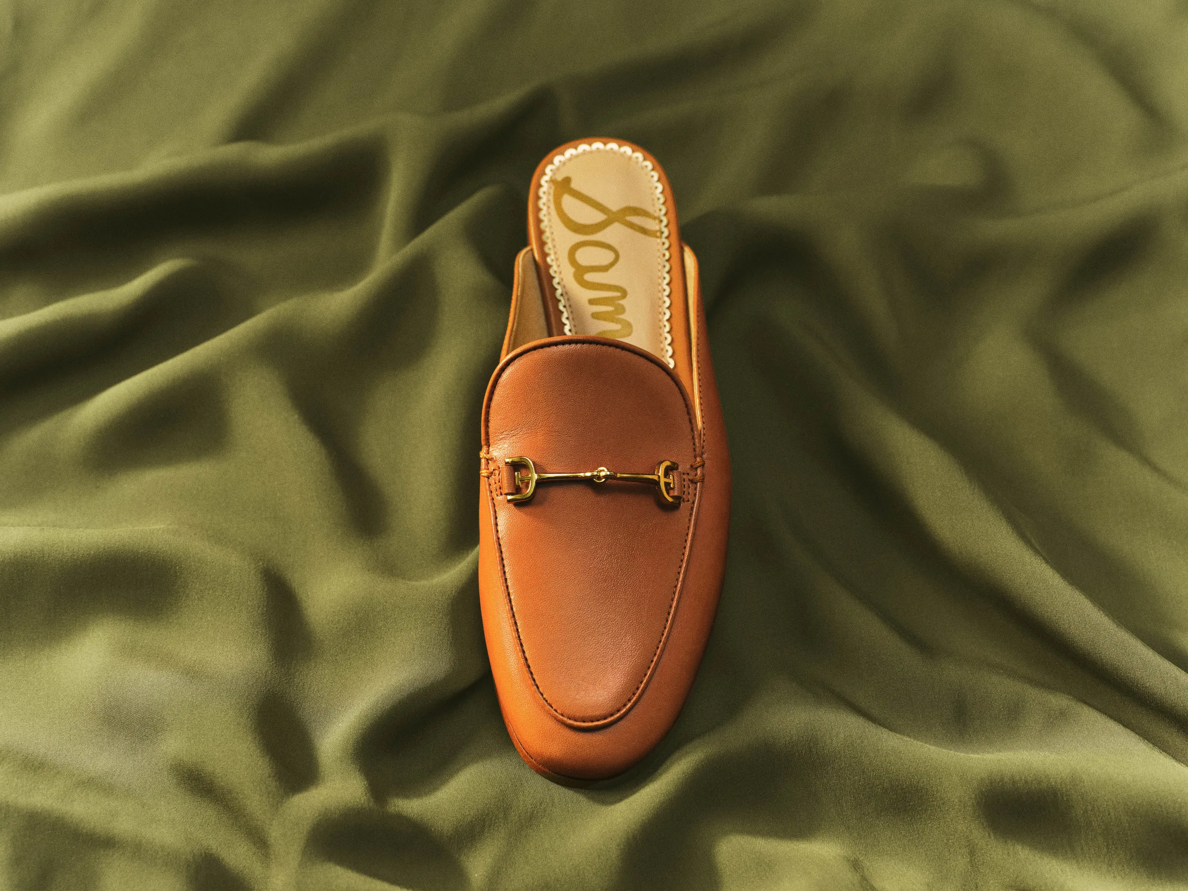 Tan leather Linnie mule with gold horsebit detail laid on rumpled olive green fabric, for Sam Edelman.