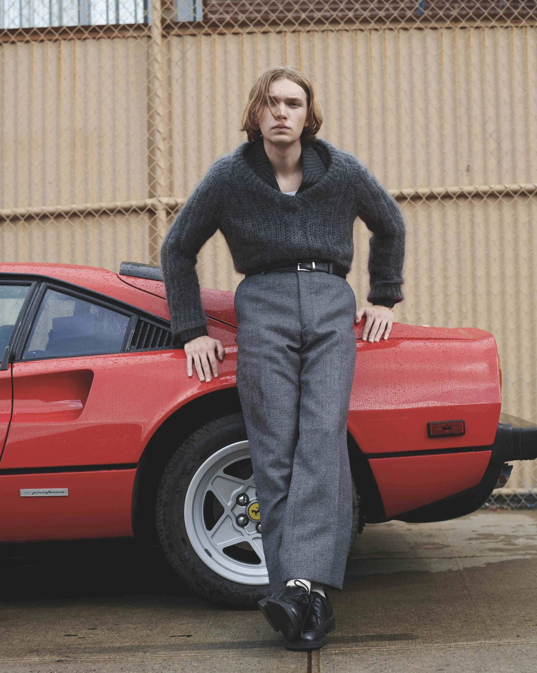 Model in grey mohair sweater and wide wool trousers leaning on a red Ferrari by a chain-link fence, for L'Officiel Hommes.