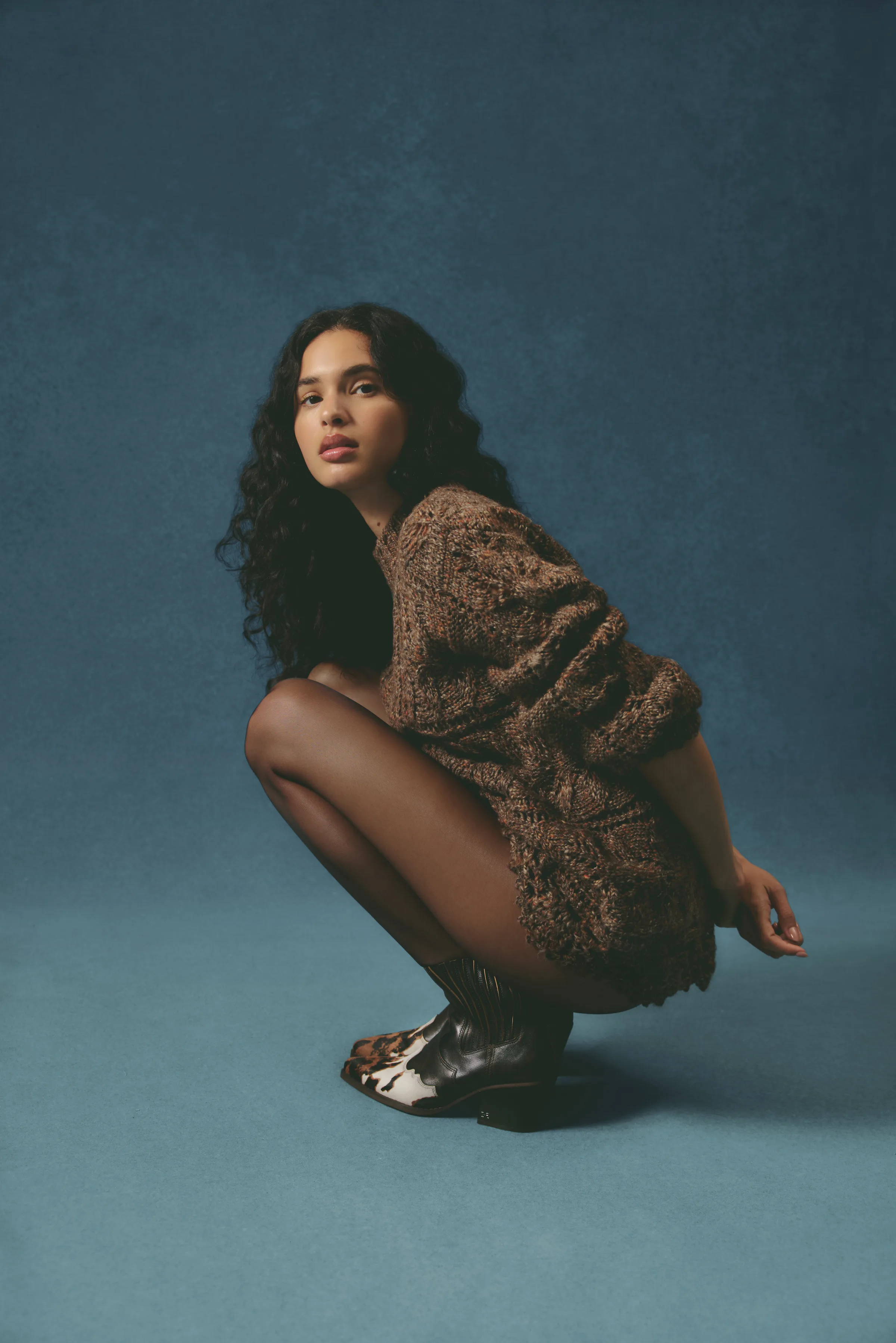 Model crouching in a brown knit sweater and brown cowhide western boots against a teal backdrop, for Sam Edelman.