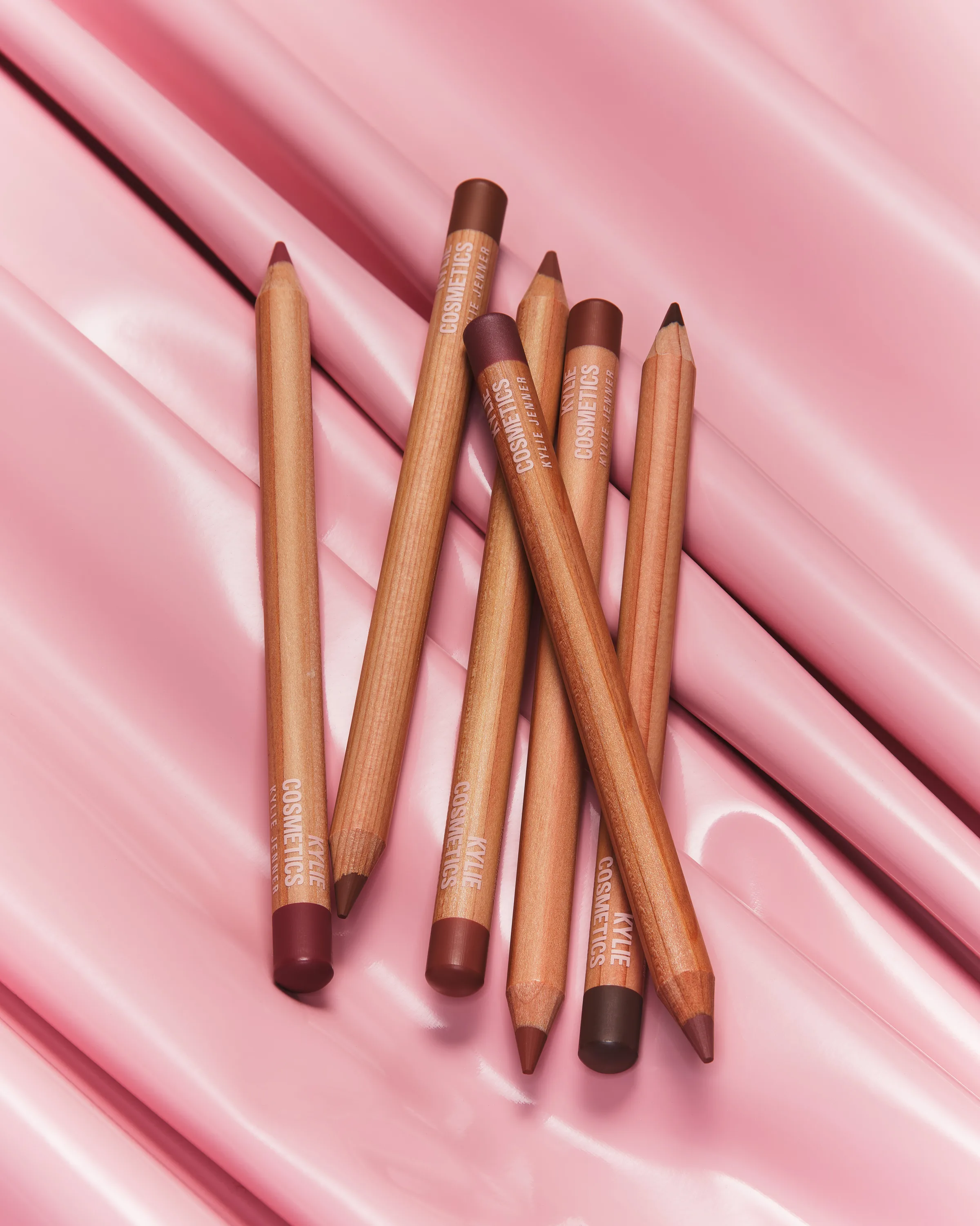 Six wooden lip liner pencils in red and brown shades lying on glossy pink satin, for Kylie Cosmetics.