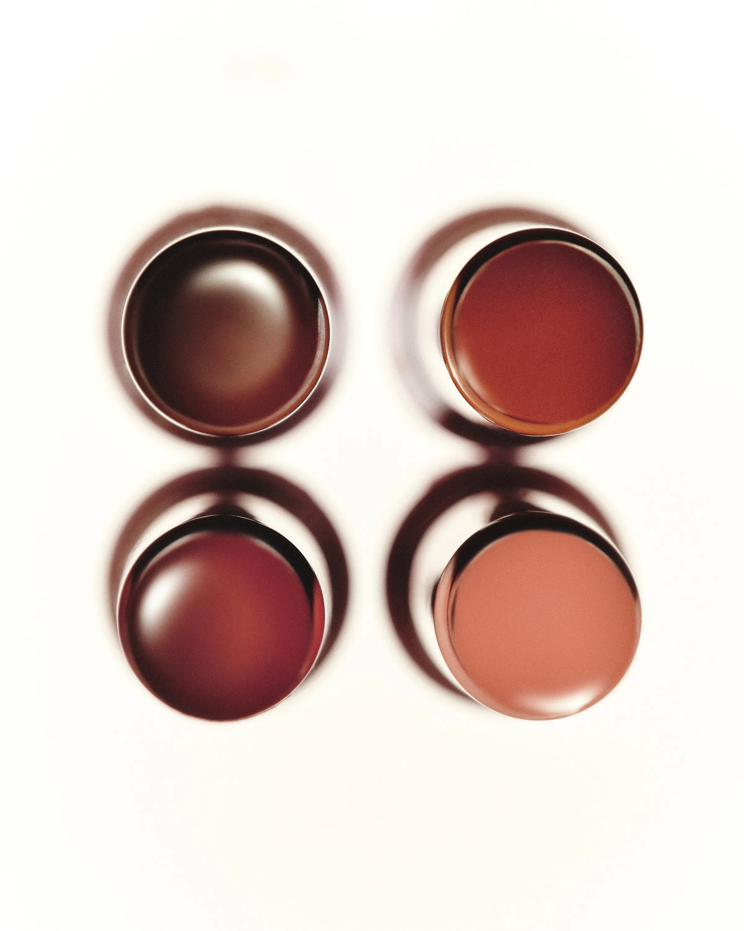 Overhead still life of four lip balms in brown, red and nude shades casting soft shadows on white, for Kylie Cosmetics.