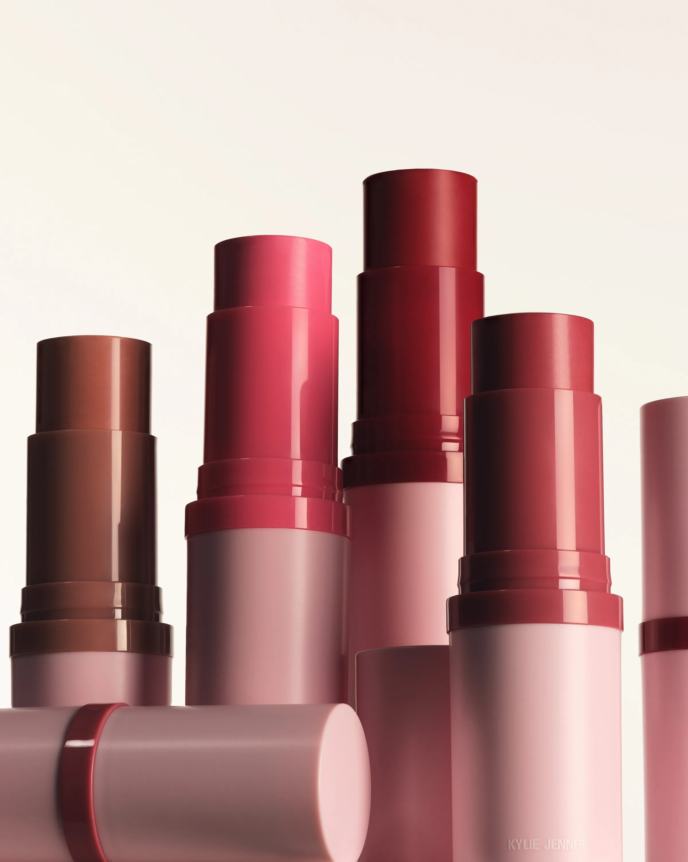 Uncapped blush sticks in brown, pink and berry shades with pink tubes on a cream background, for Kylie Cosmetics.