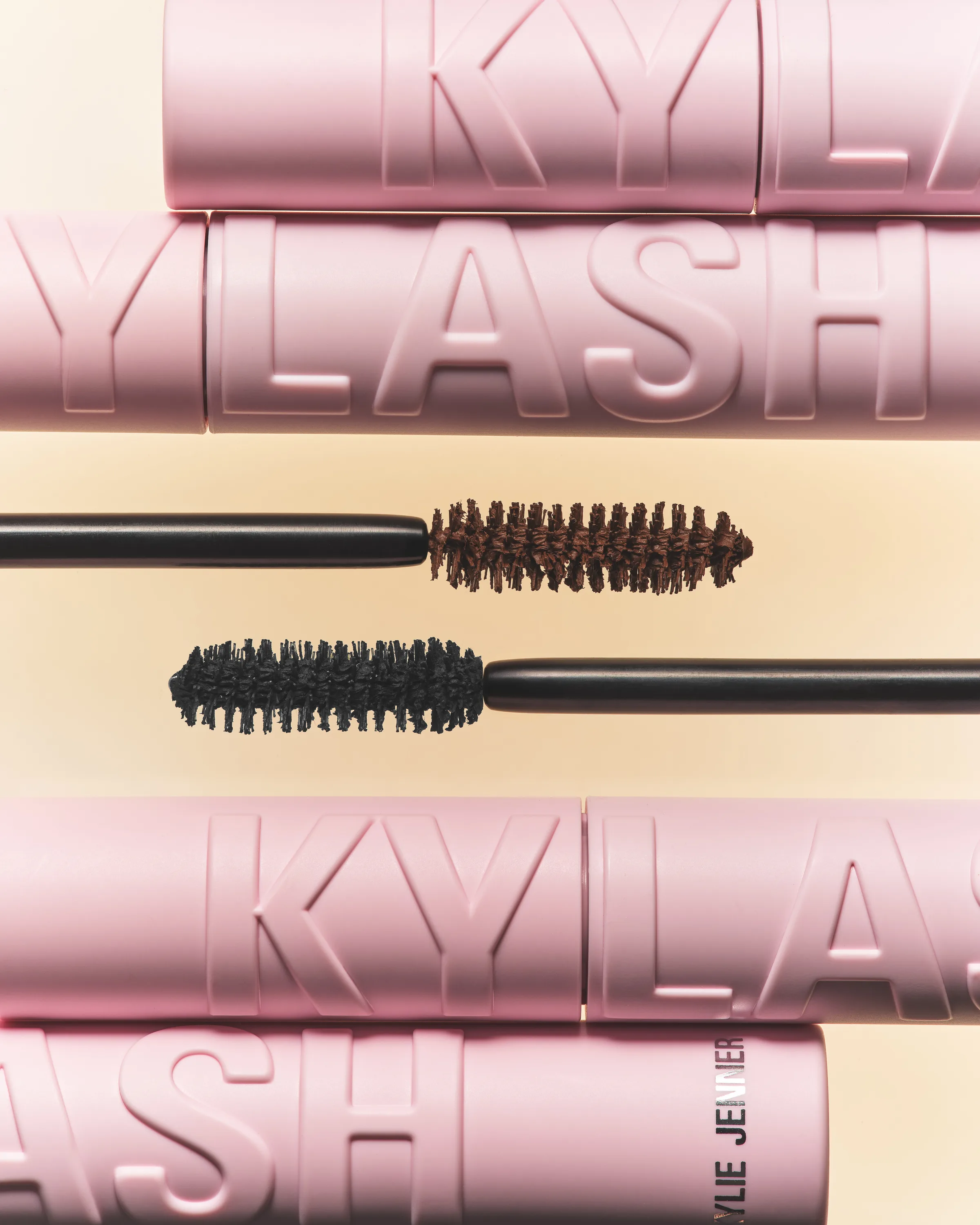 Brown and black mascara wands floating between stacked pink Kylash tubes on a peach background, for Kylie Cosmetics.