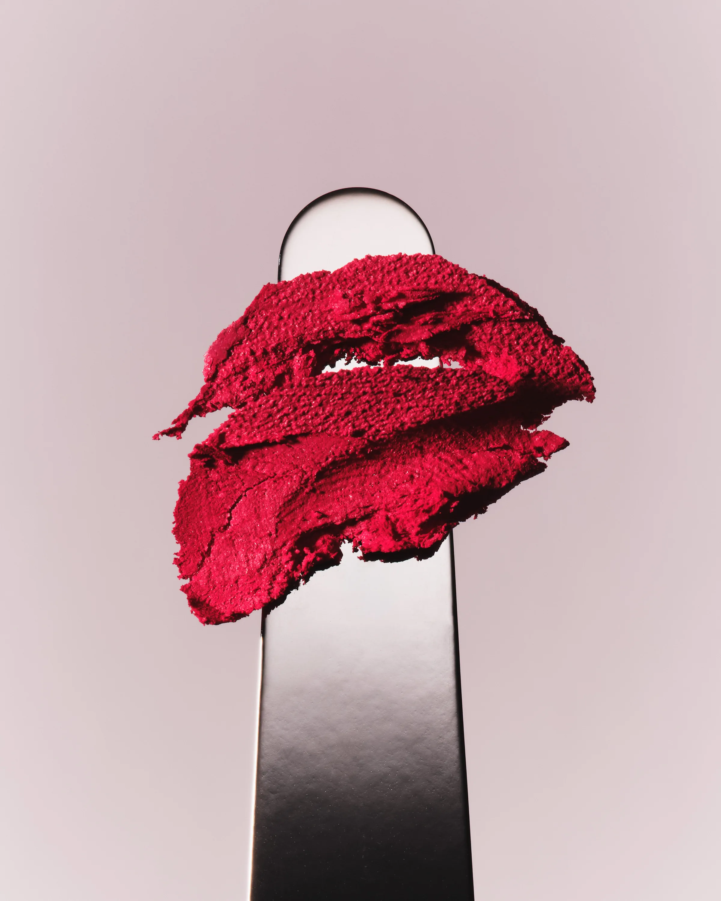 Red lipstick smear on a metal spatula against a pale pink-grey background, shade Lady Bug, for Kylie Cosmetics.