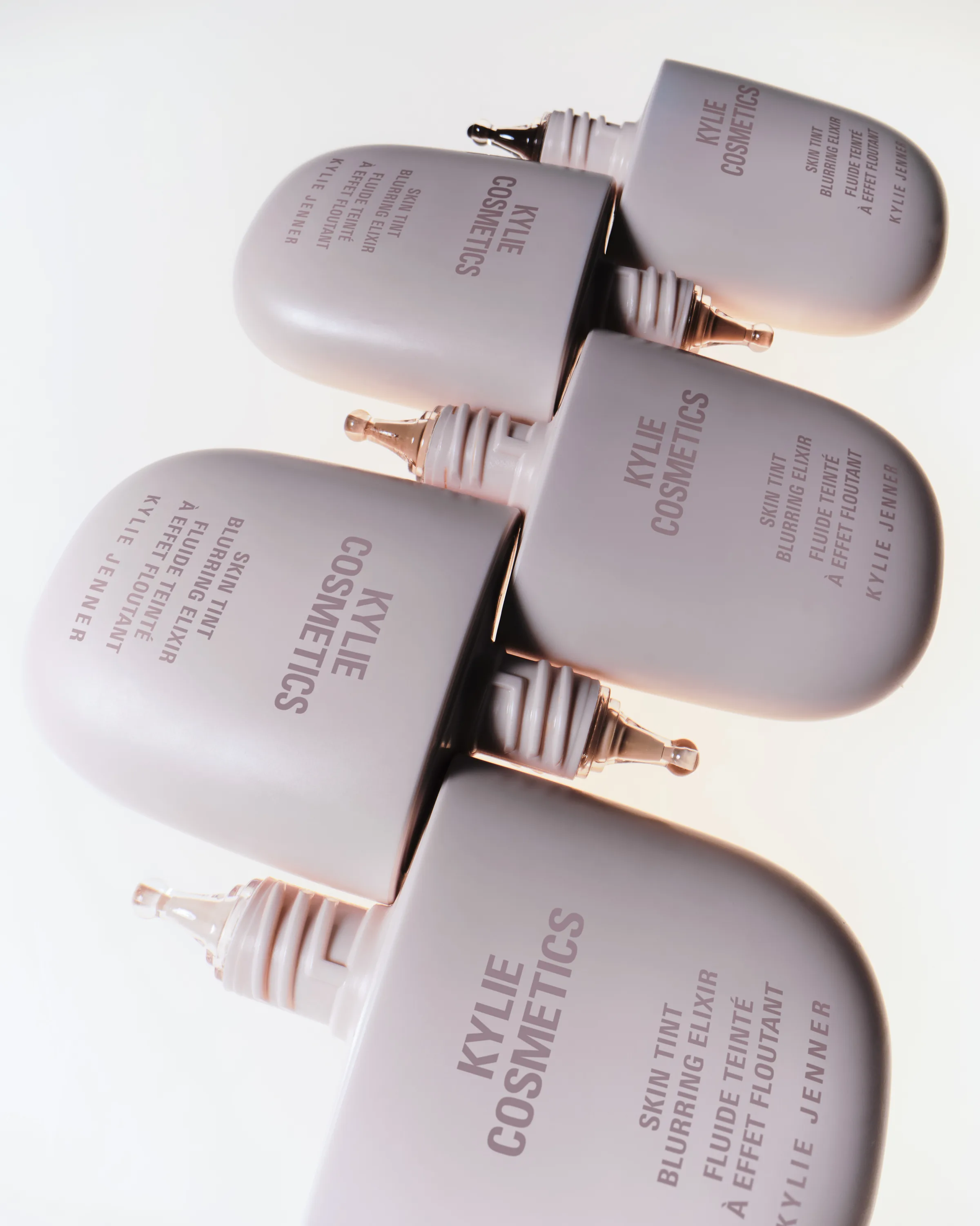 Five pale pink Skin Tint Blurring Elixir bottles stacked diagonally with dropper tips on a white background, for Kylie Cosmetics.