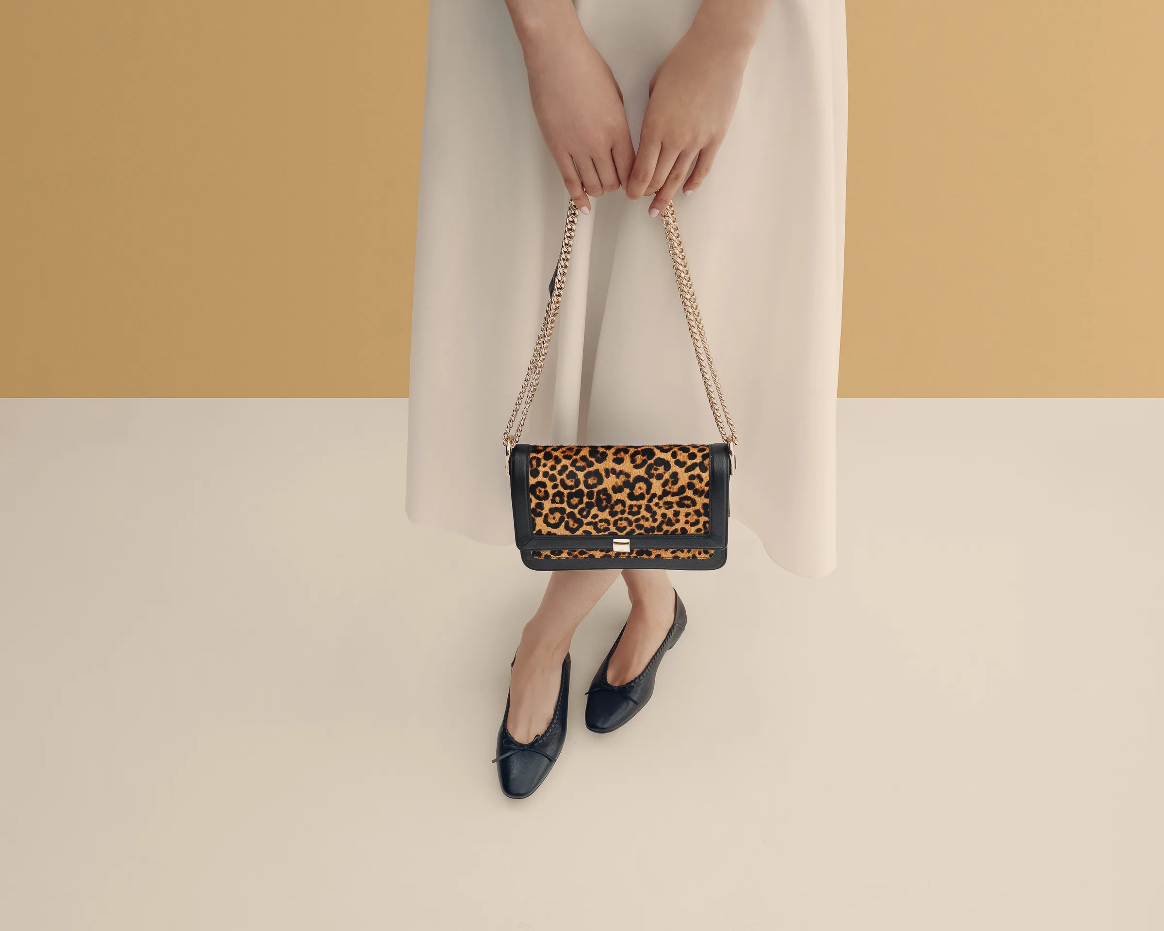 Model in cream skirt holding a leopard calf hair chain shoulder bag, wearing black ballet flats, on a tan set, for Cole Haan.