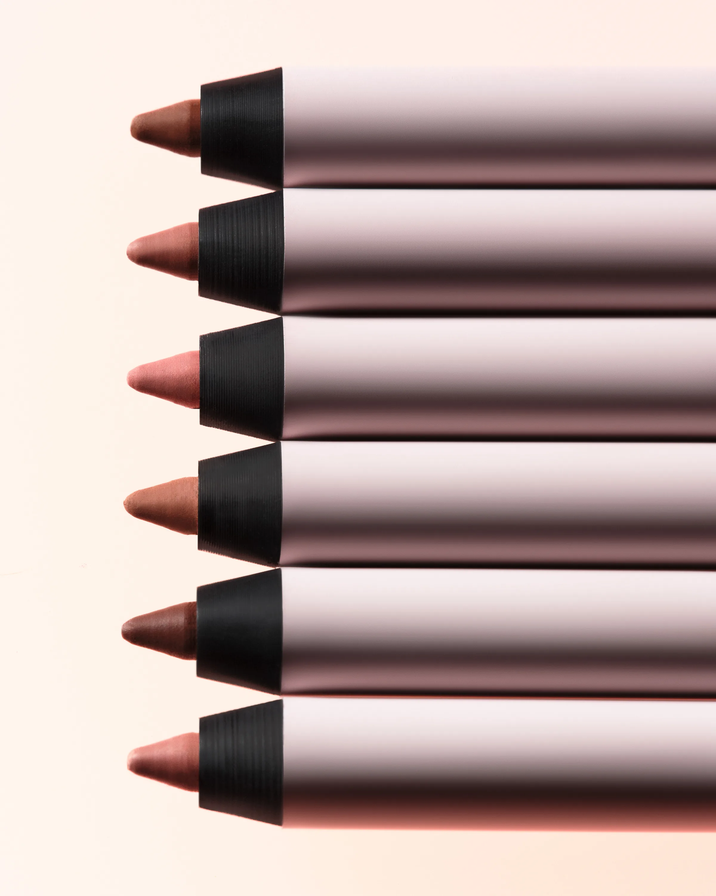 Six nude and brown lip liner pencils with black tips lined up on a soft peach background, for Kylie Cosmetics.