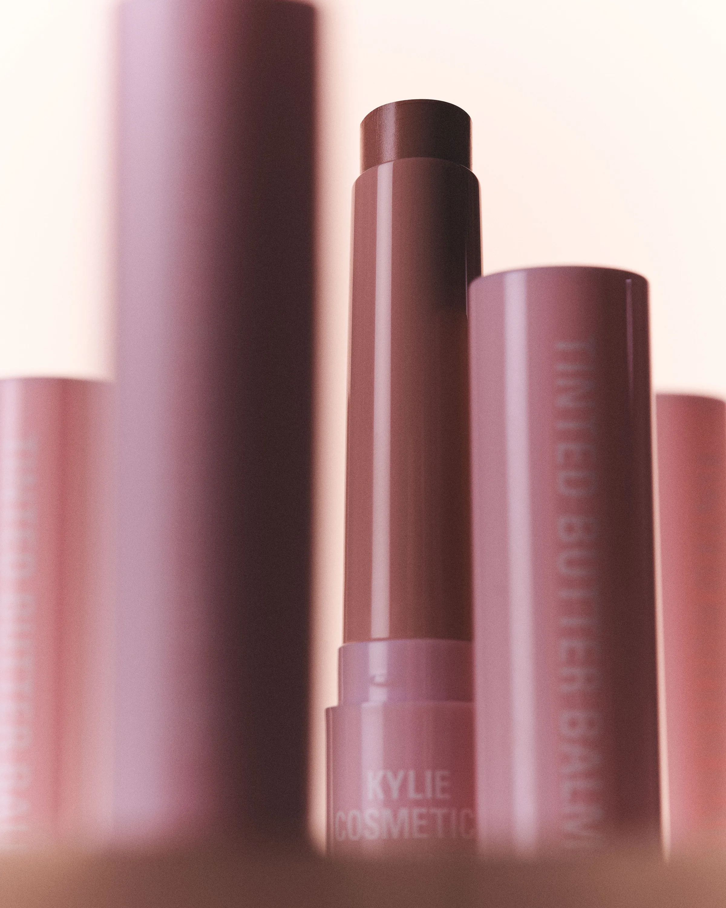 Uncapped Tinted Butter Balm lipstick in Angel Cake among pink tubes on a soft pink background, for Kylie Cosmetics.