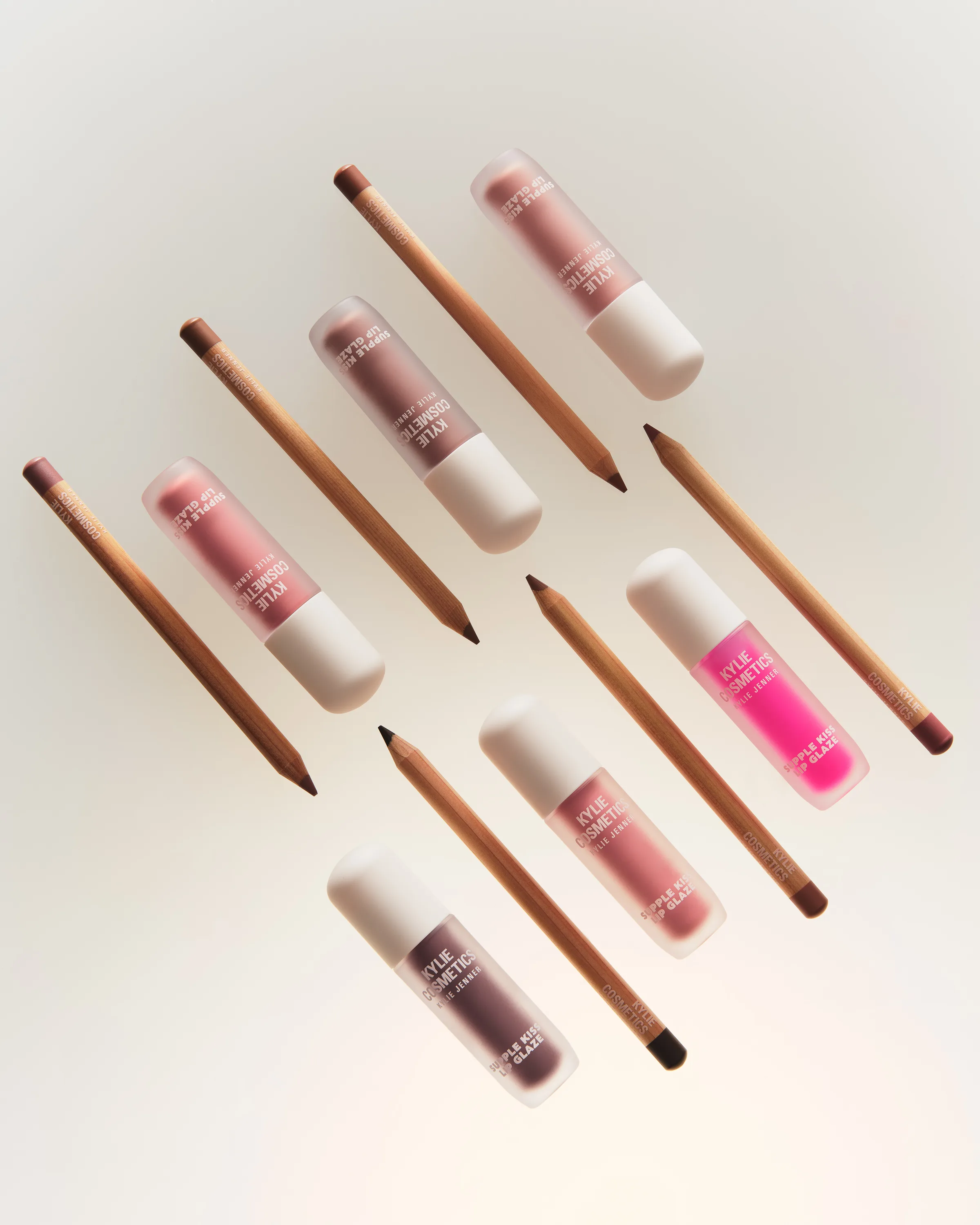 Pink and mauve lip glazes with wooden lip liners arranged diagonally on a soft cream background, for Kylie Cosmetics.