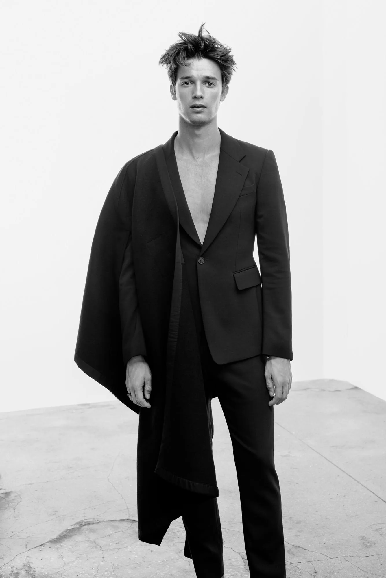 Black-and-white portrait of a shirtless man in a black suit with a draped cape panel on a concrete floor, for Man About Town.