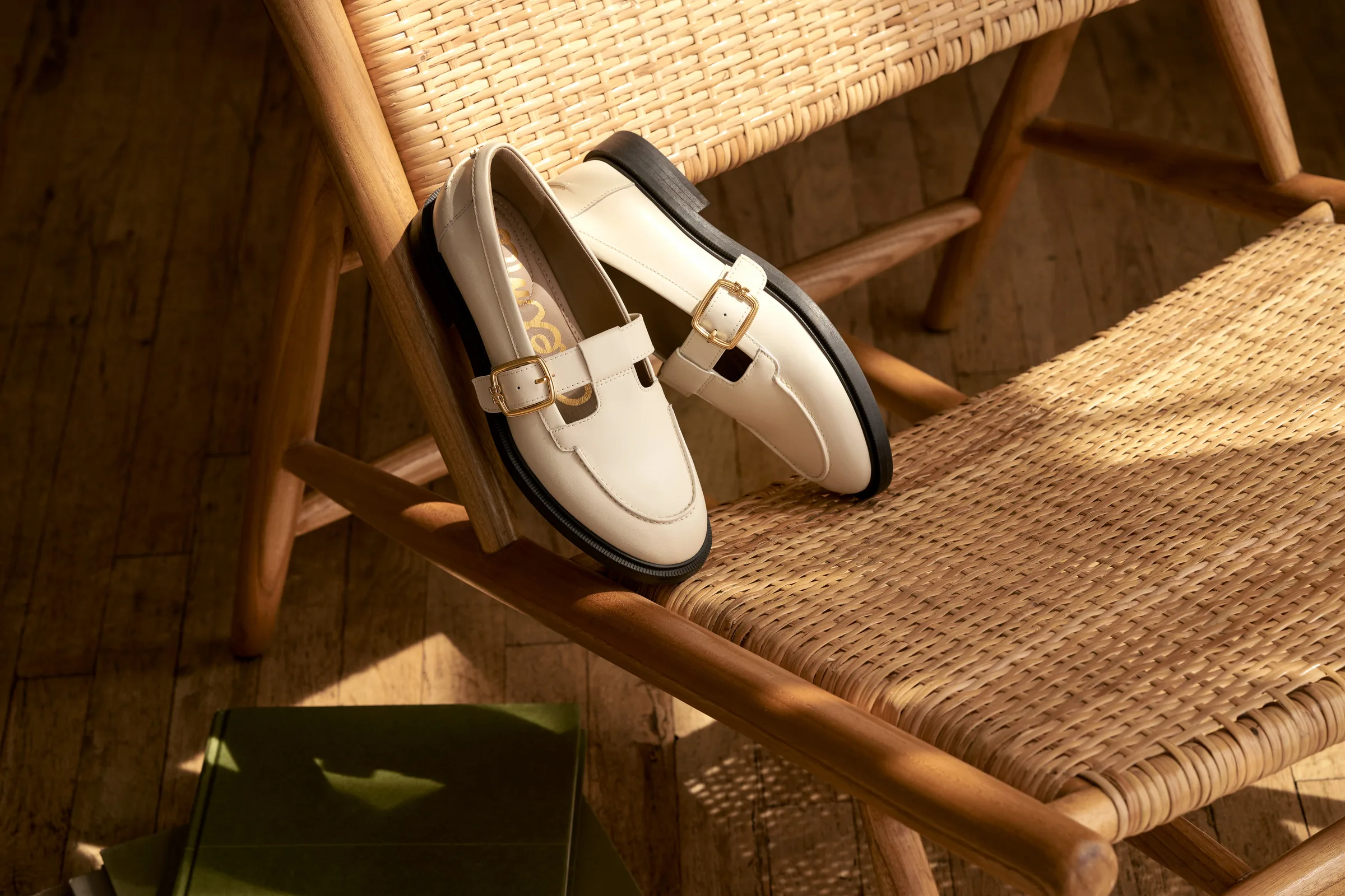 Cream leather loafers with gold buckles resting on a sunlit rattan chair beside a green shoebox, for Sam Edelman.
