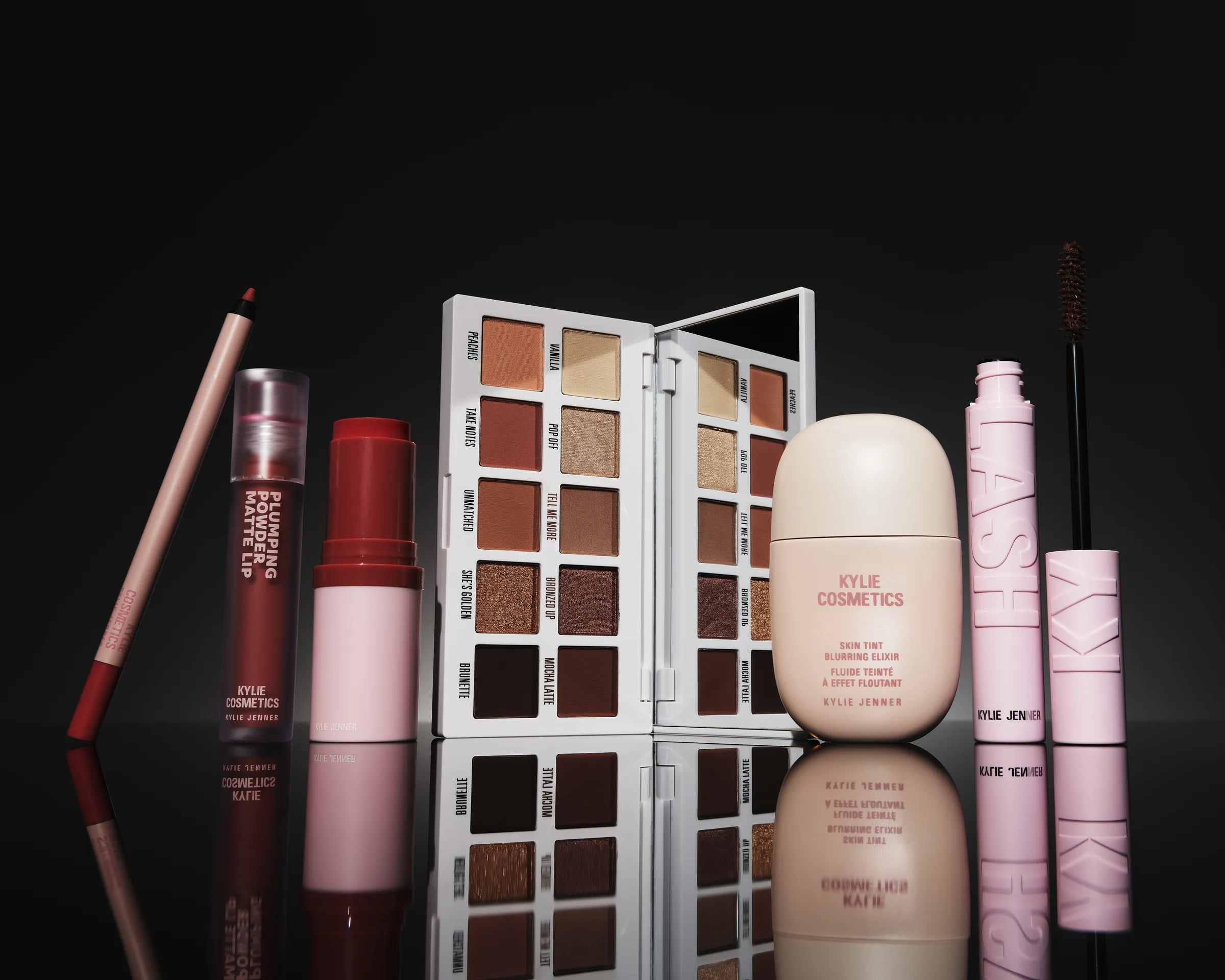 Still life of lip pencil, matte lip, blush stick, bronze palette, skin tint and mascara on reflective black, for Kylie Cosmetics.