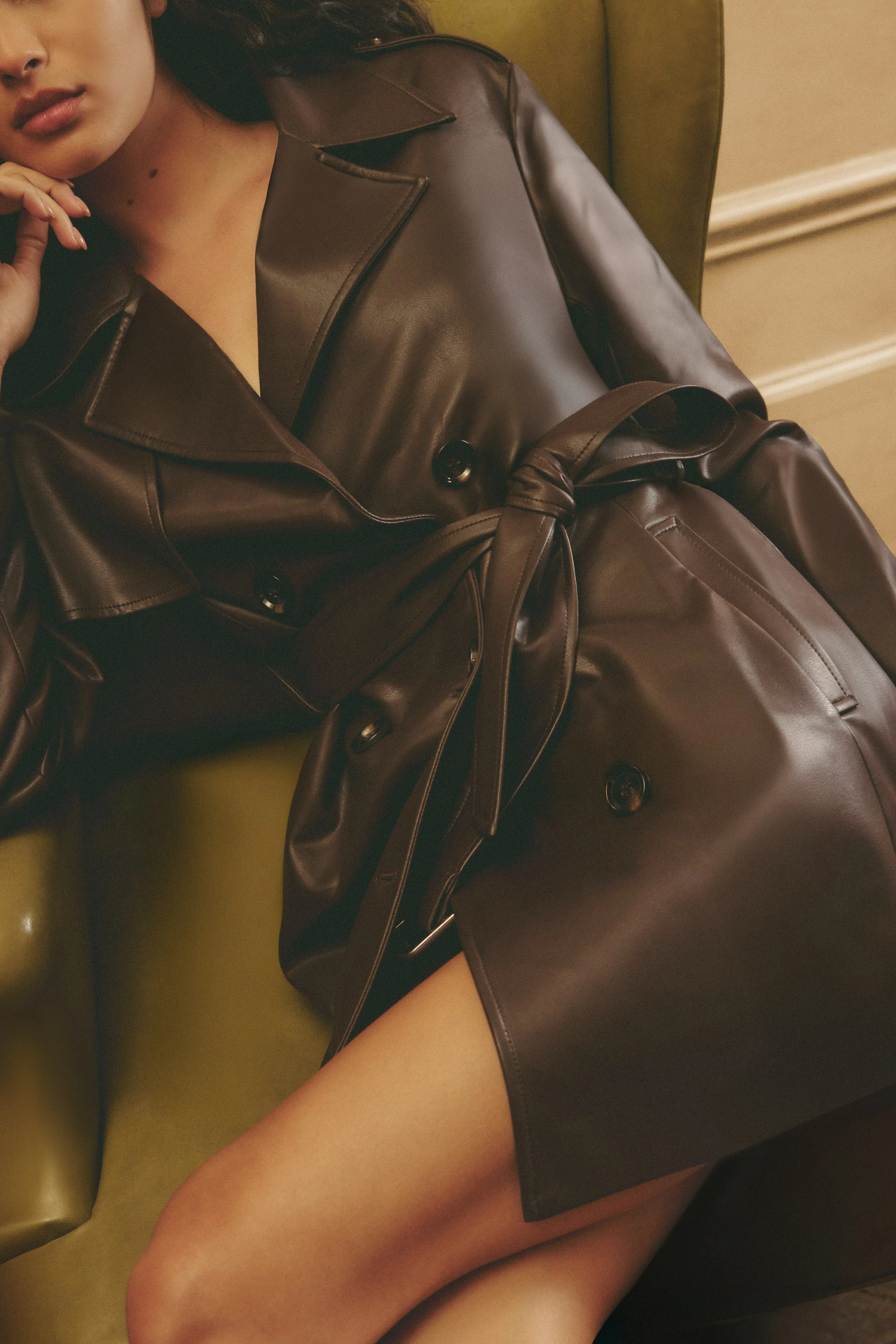 Model in a belted brown leather trench coat lounging on an olive green armchair, for Sam Edelman.