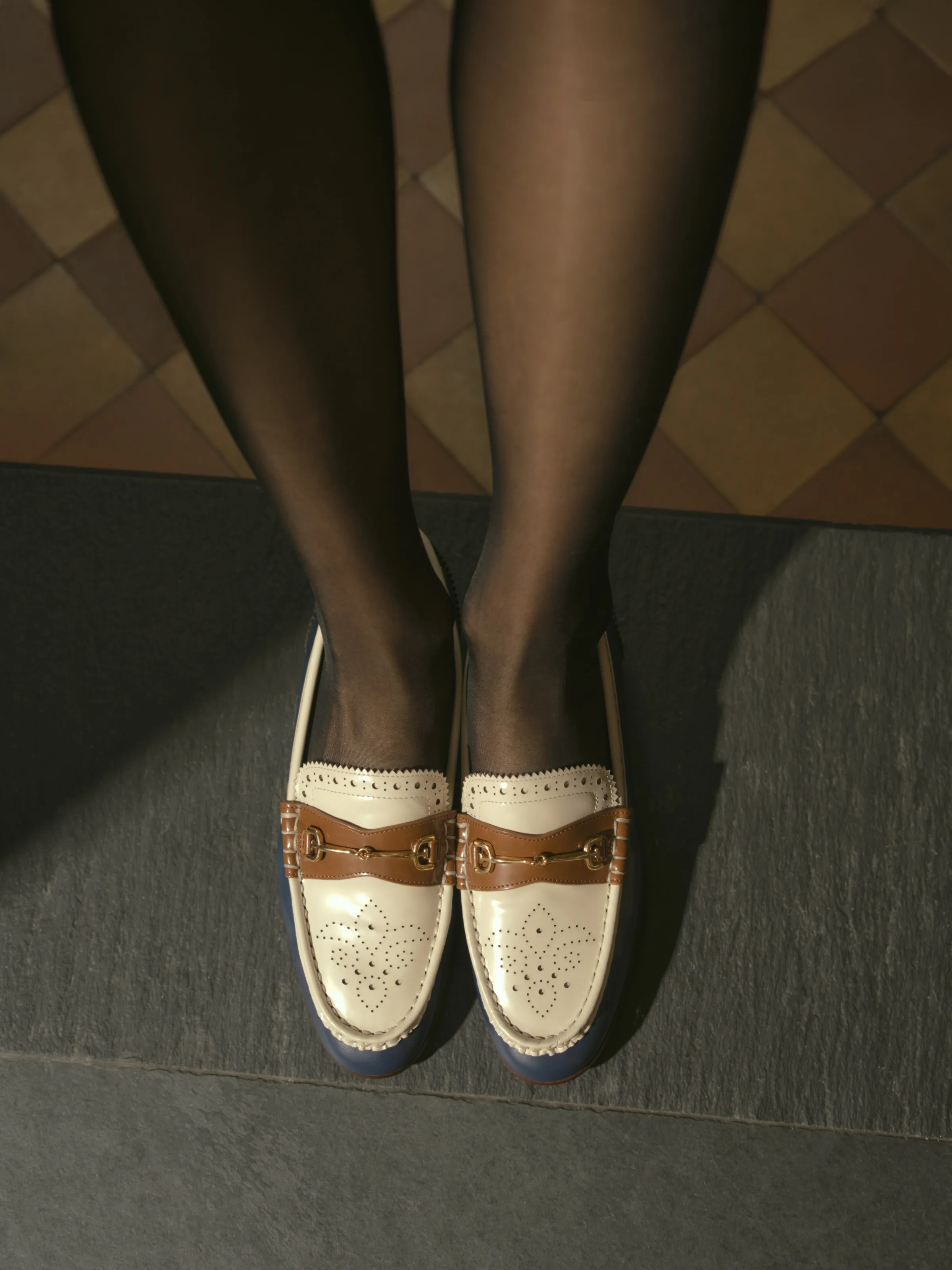 Legs in sheer black tights wearing cream patent brogue loafers with tan horsebit strap on grey stone step, for Sam Edelman.