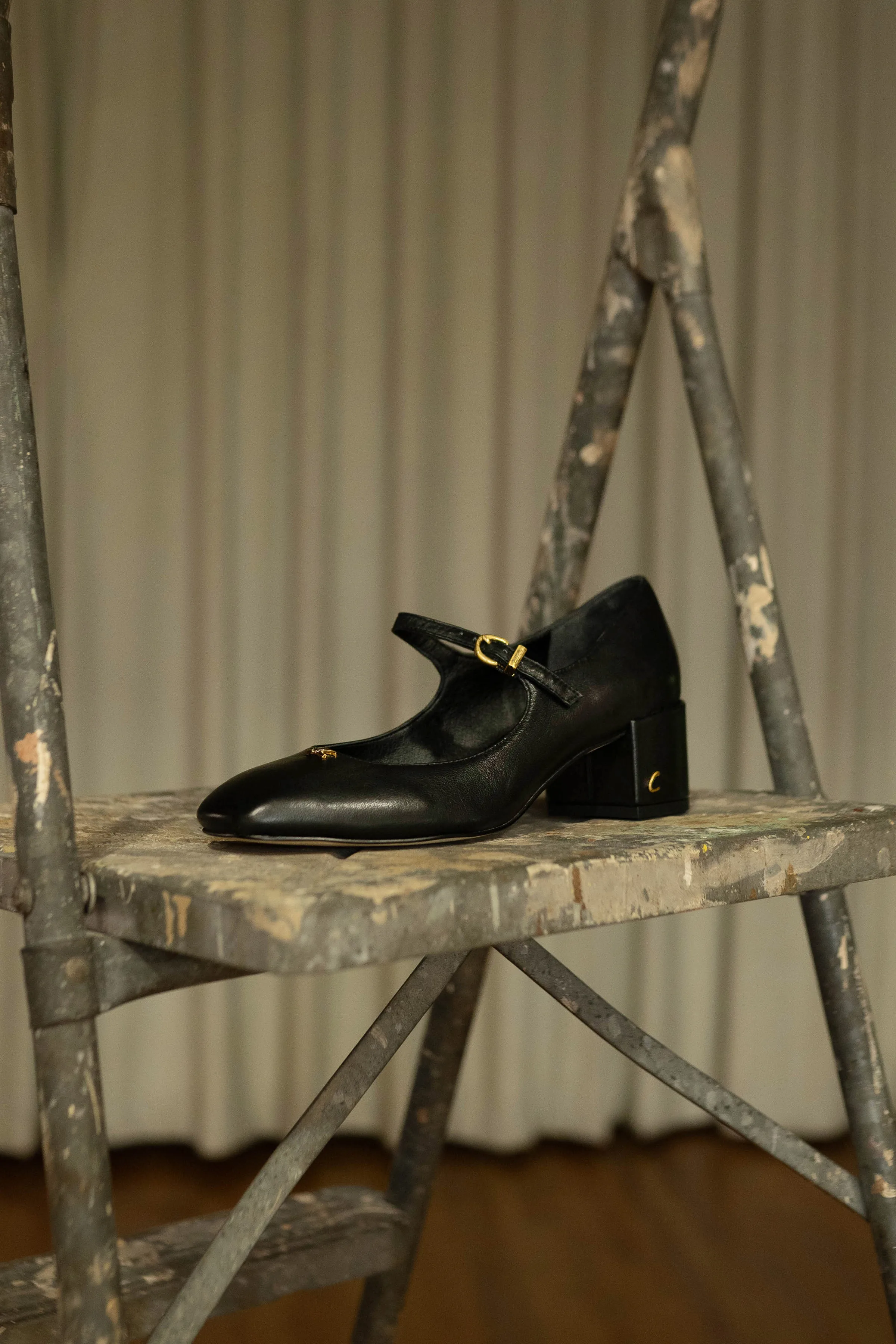 Black leather Mary Jane heel with gold buckle on a paint-splattered metal stepladder before a curtain, for Sam Edelman.