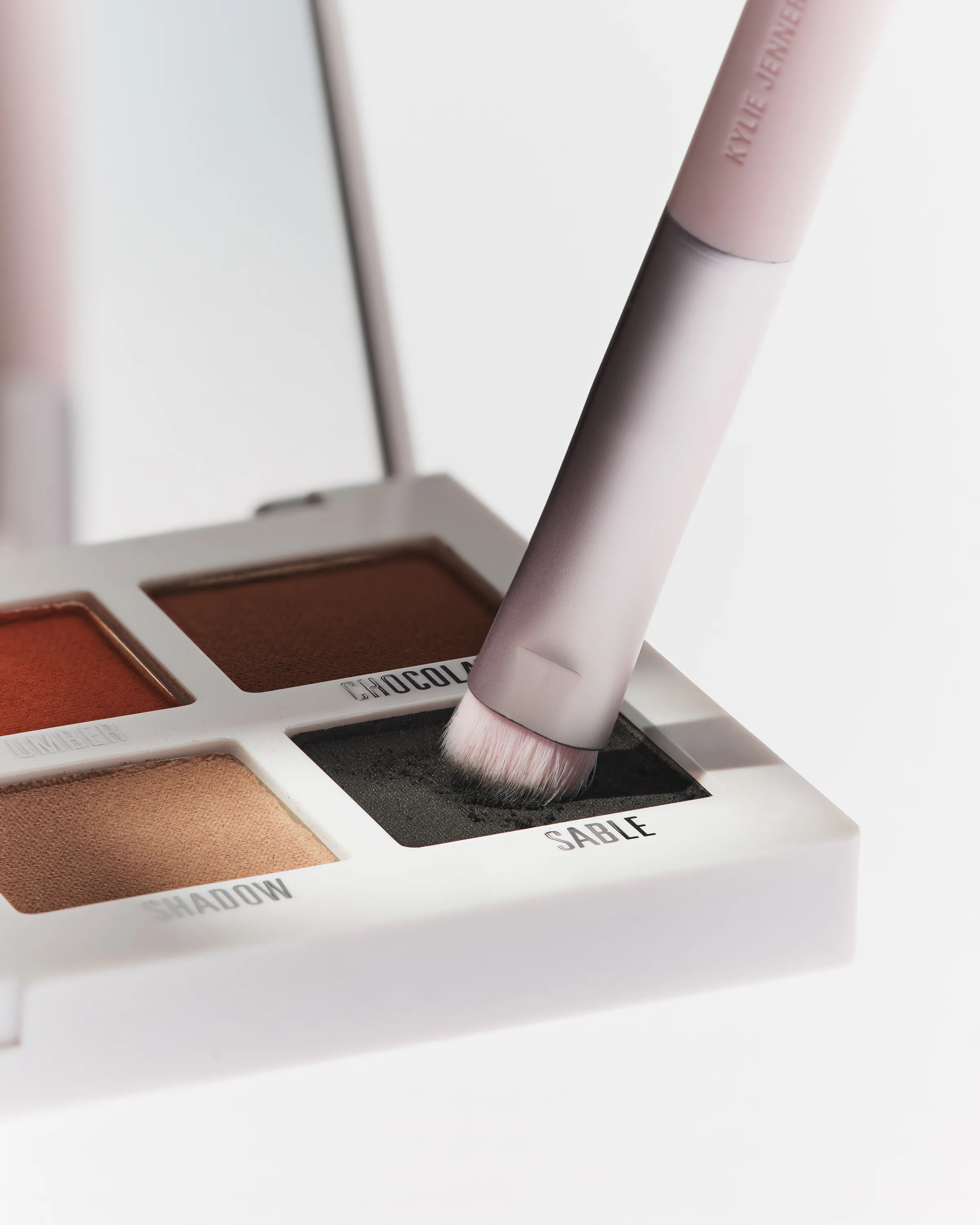 Pink eyeshadow brush dipping into the dark Sable shade of a white quad palette with mirror, for Kylie Cosmetics.