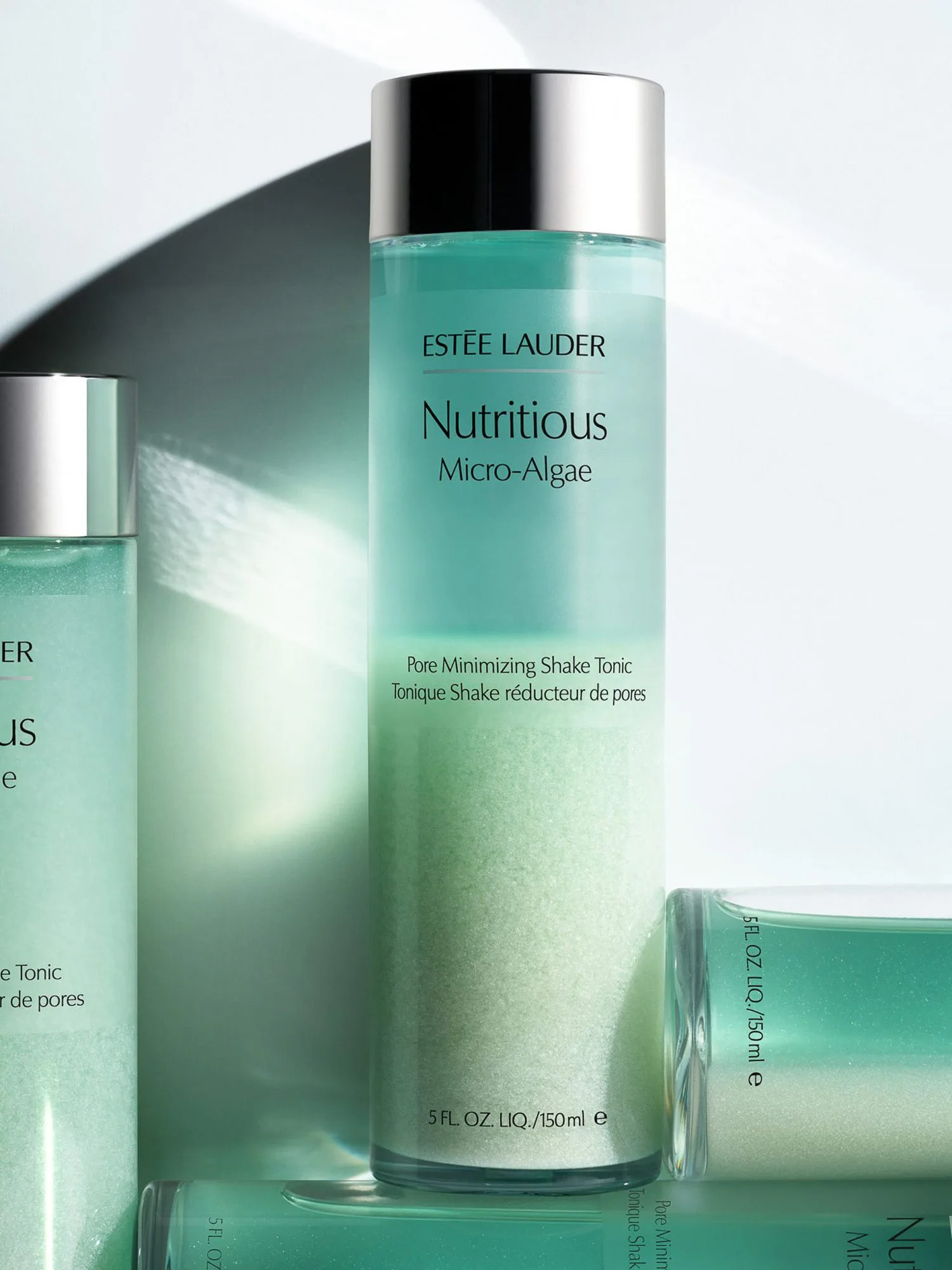 Mint-green Nutritious Micro-Algae tonic bottles with silver caps in sunlight and shadow on white, for Estée Lauder.