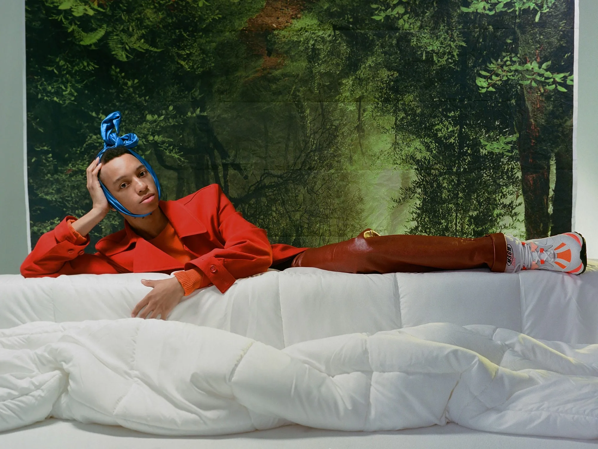 Model in red coat and blue headscarf reclining on a white duvet before a forest backdrop, for VMAN.