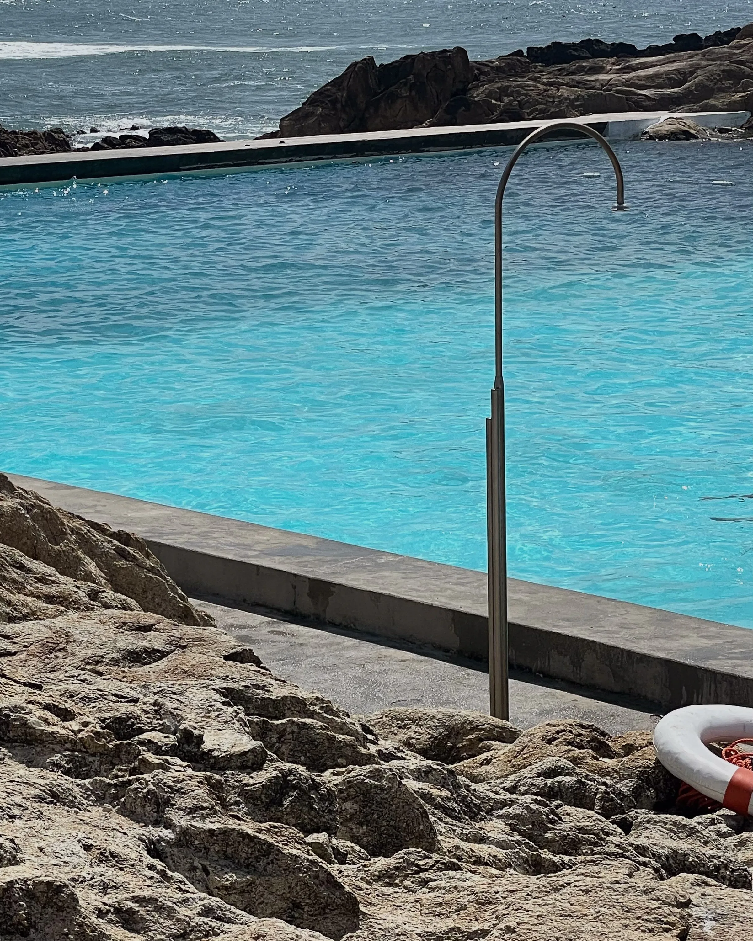 Steel outdoor shower beside a turquoise seawater pool with rocks, ocean and a red-and-white life ring.