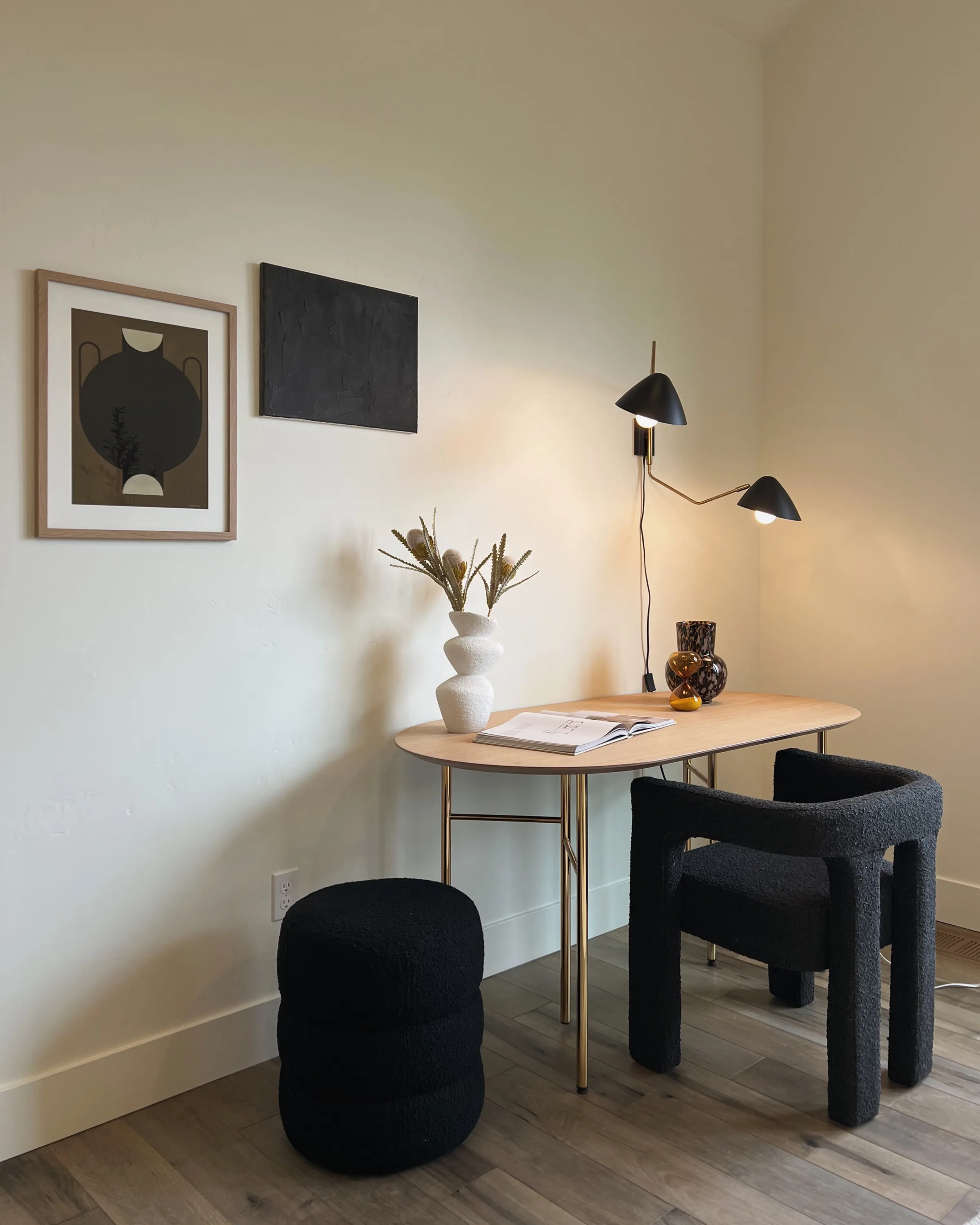Oval wood desk with black bouclé chair and pouf, sculptural white vase and black wall lamp in a sunlit corner.