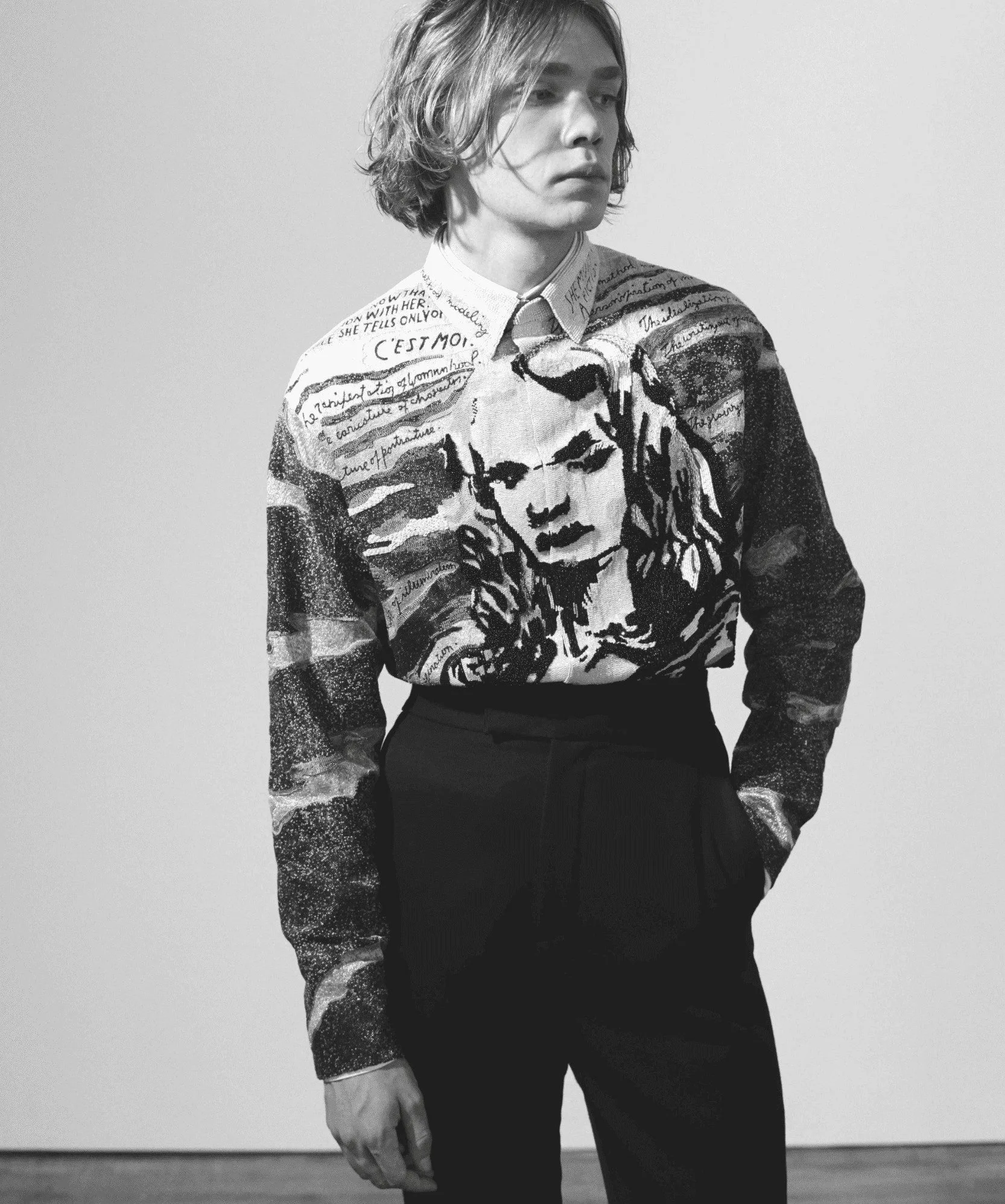 Black and white portrait of a model in an embroidered portrait-print shirt and black trousers, for L'Officiel Hommes.