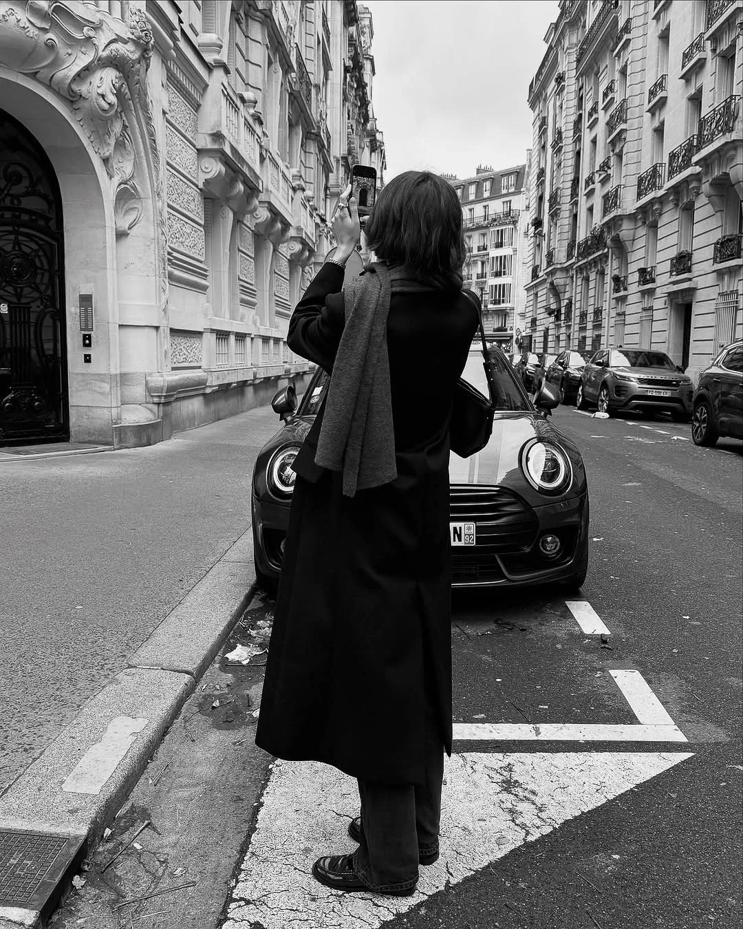Black-and-white shot of a woman in a long black coat and grey scarf taking a phone photo on a Parisian street.
