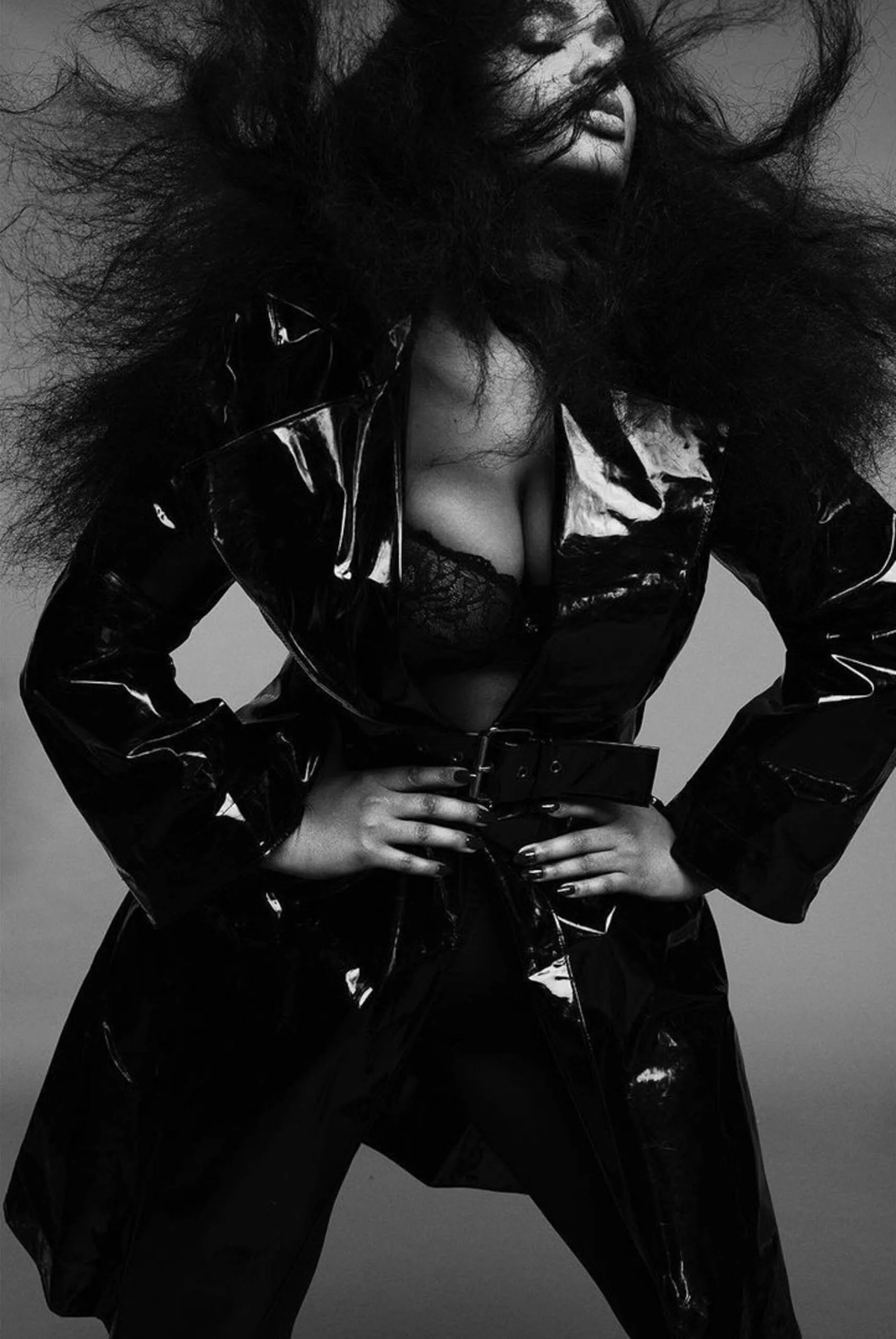 Black and white shot of a model in a glossy black patent trench coat and lace bra, hair blown wild, for V Magazine.