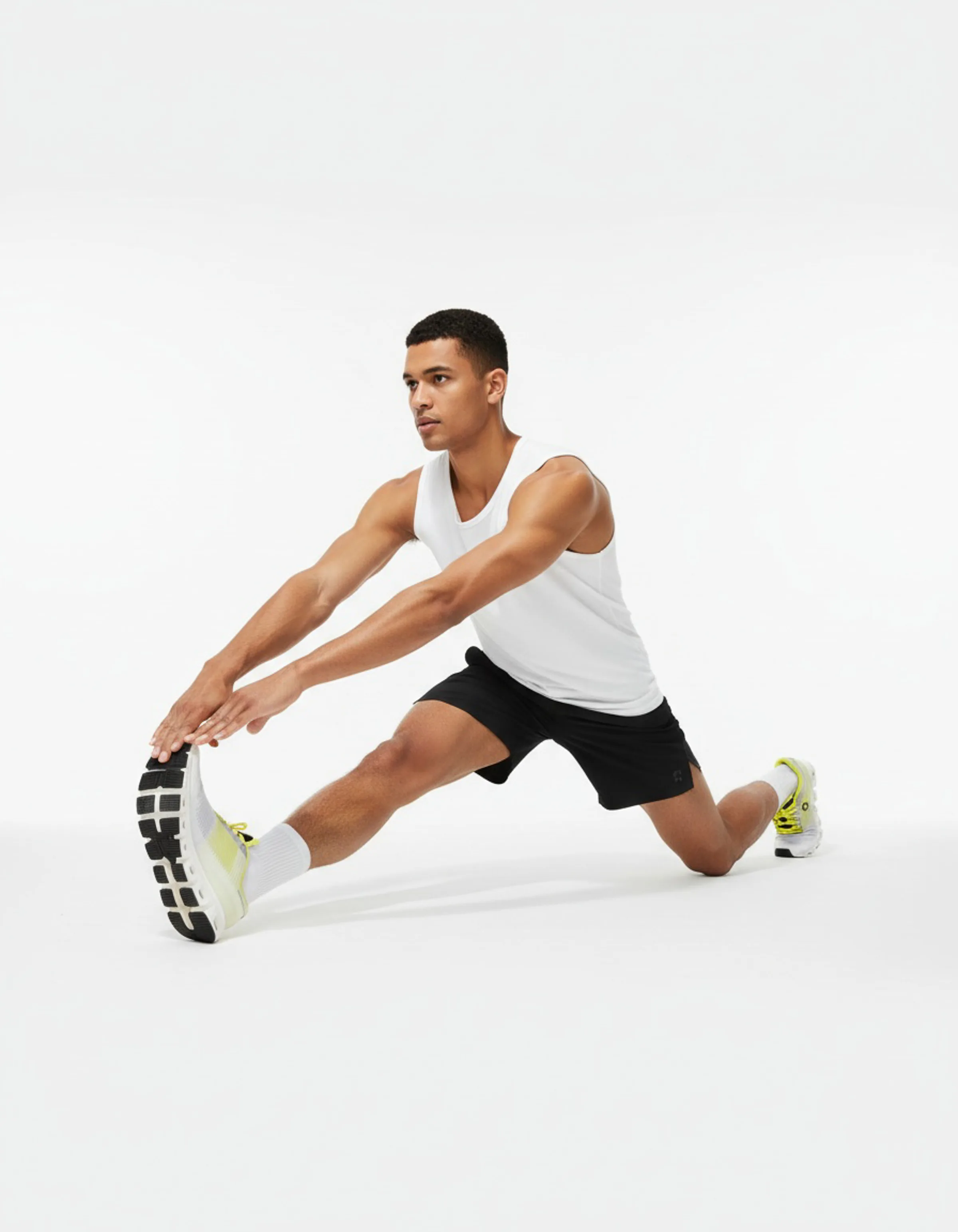 Man in white tank top and black shorts stretching his hamstring in white and lime running shoes on a white backdrop. On AI Concept.