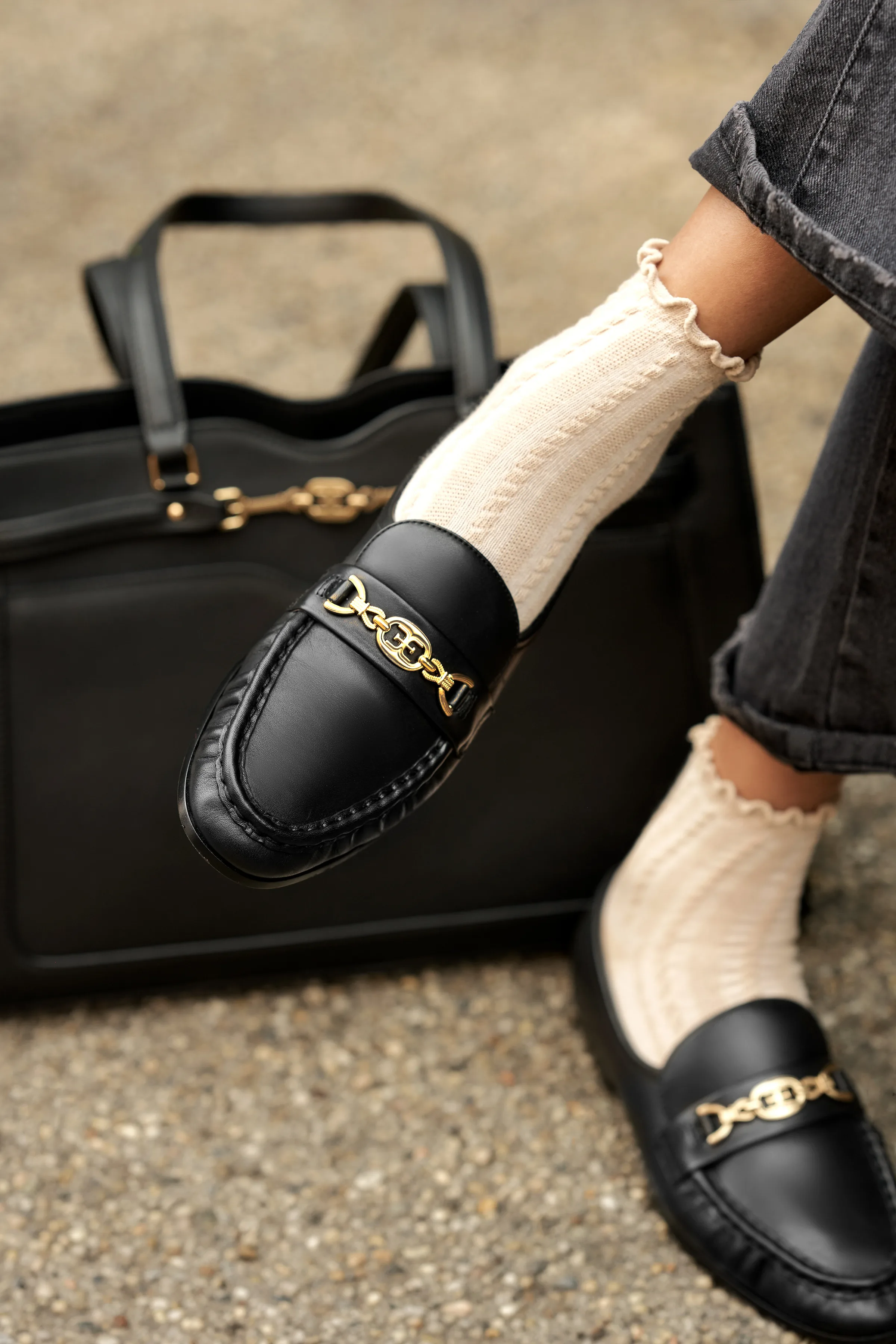 Black leather loafers with gold chain bits worn with cream ruffle socks beside a black tote on gravel, for Sam Edelman.