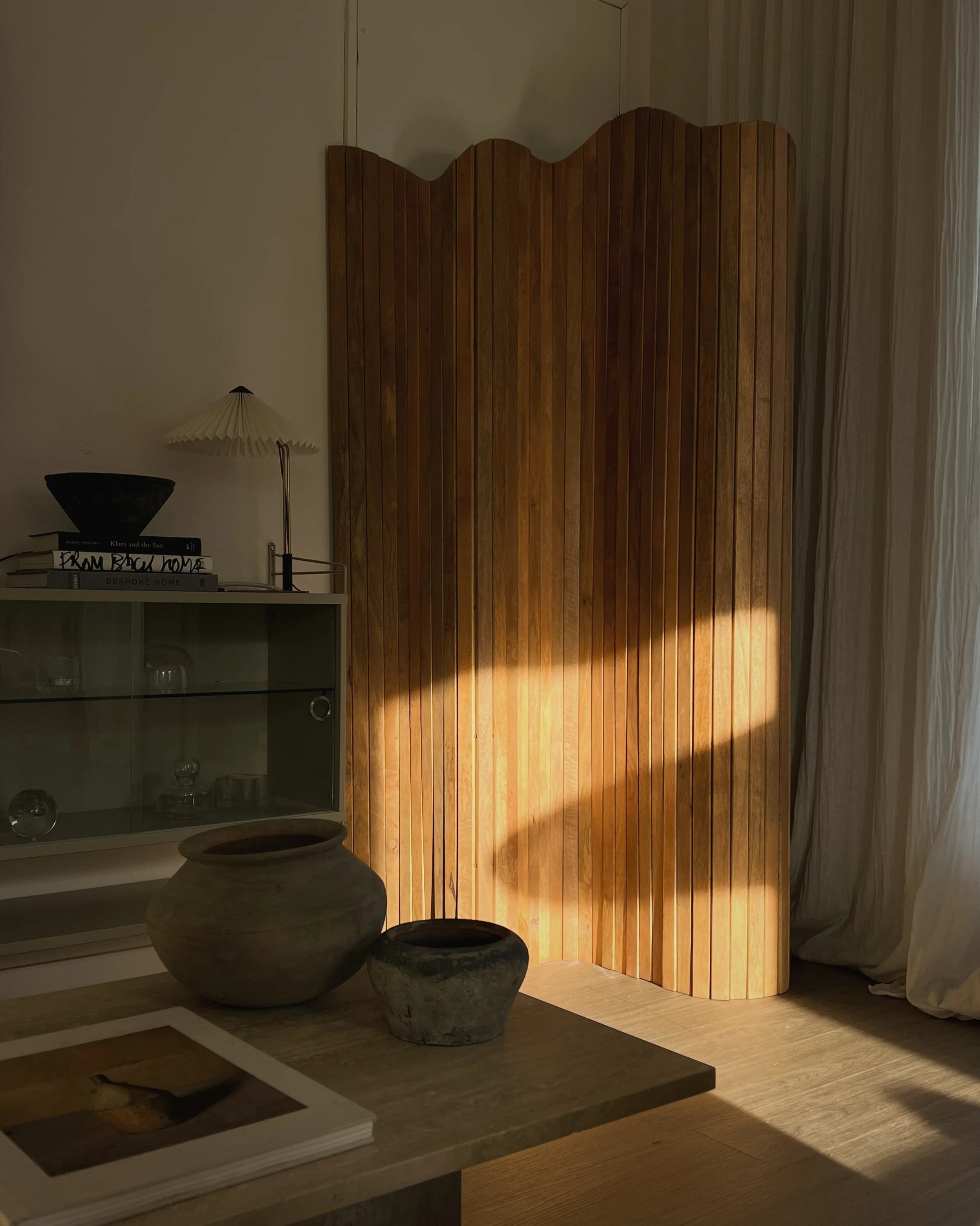 Wavy slatted wood folding screen in warm sunlight beside a glass cabinet, stone vessels and a pleated lamp, for Jennifer Hagler.