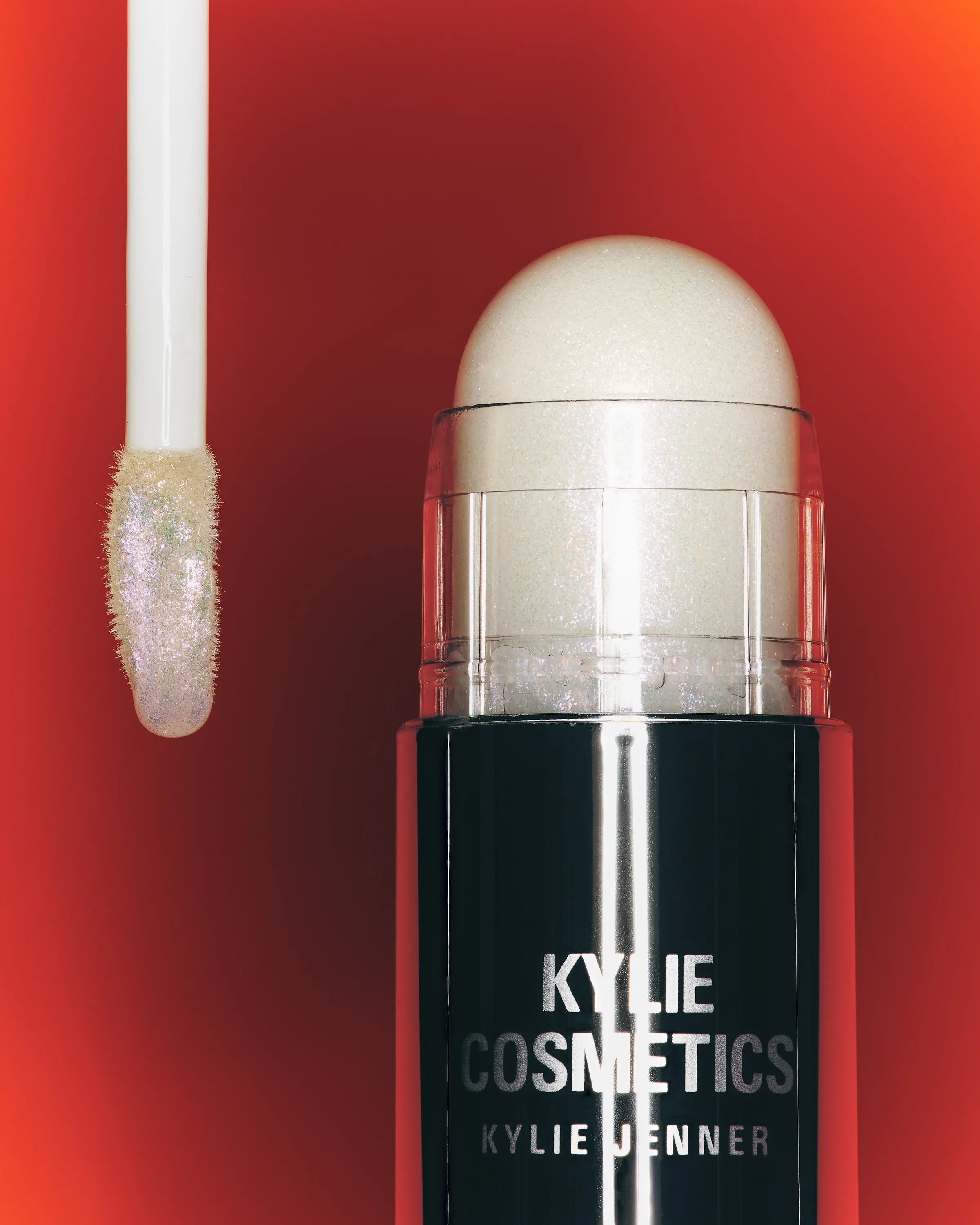 Shimmering Pink Ice lip glaze wand beside a Solar Glow dewy balm stick on a red-orange gradient, for Kylie Cosmetics.