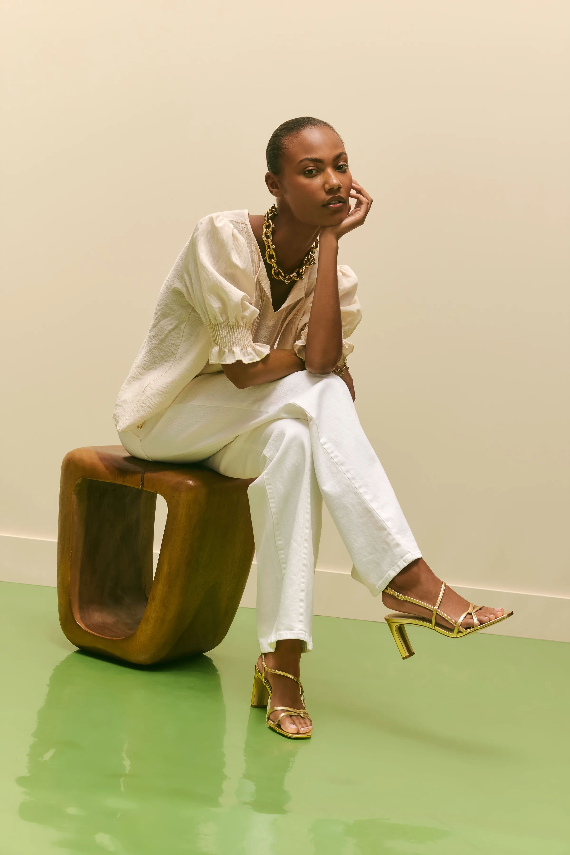 Model in white puff-sleeve top and jeans sits on a wooden stool wearing gold strappy heeled sandals, for Sam Edelman.