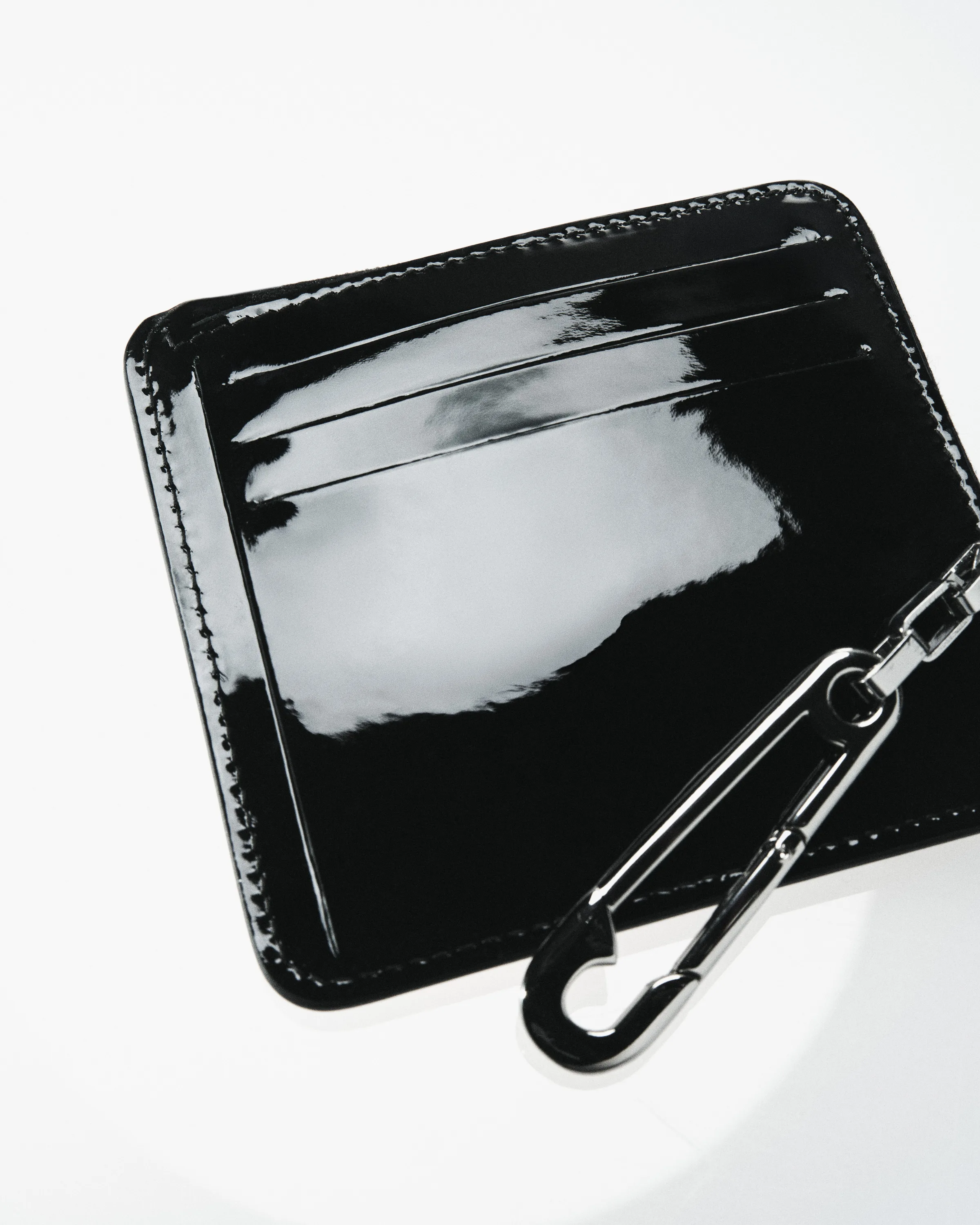 Black patent leather card wallet with silver clip hook on a white surface, for Strata.