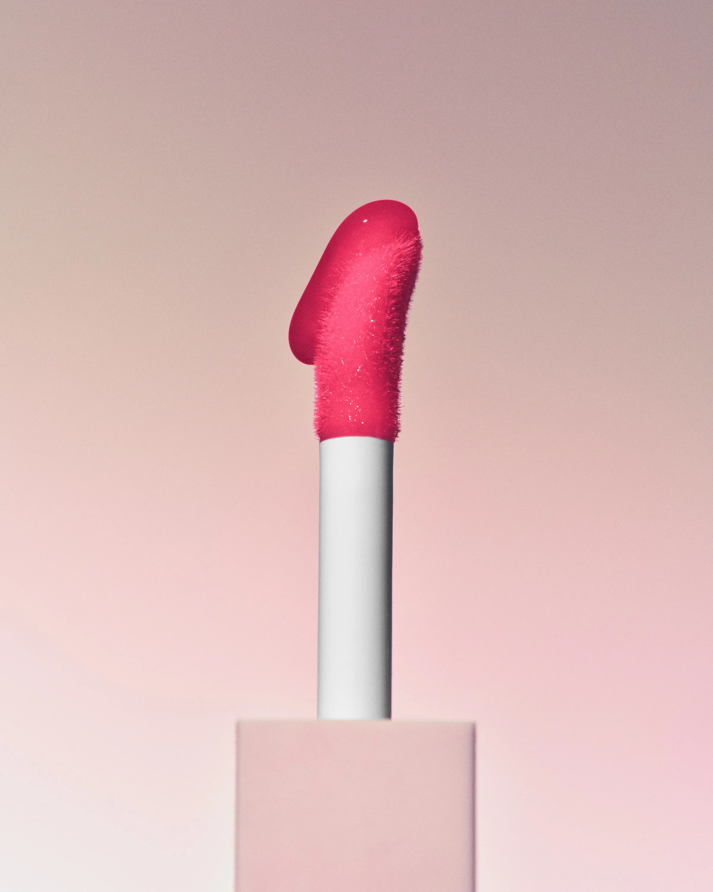 Lip applicator coated in hot pink gloss rising from a pale pink box against a soft pink gradient, for Kylie Cosmetics.