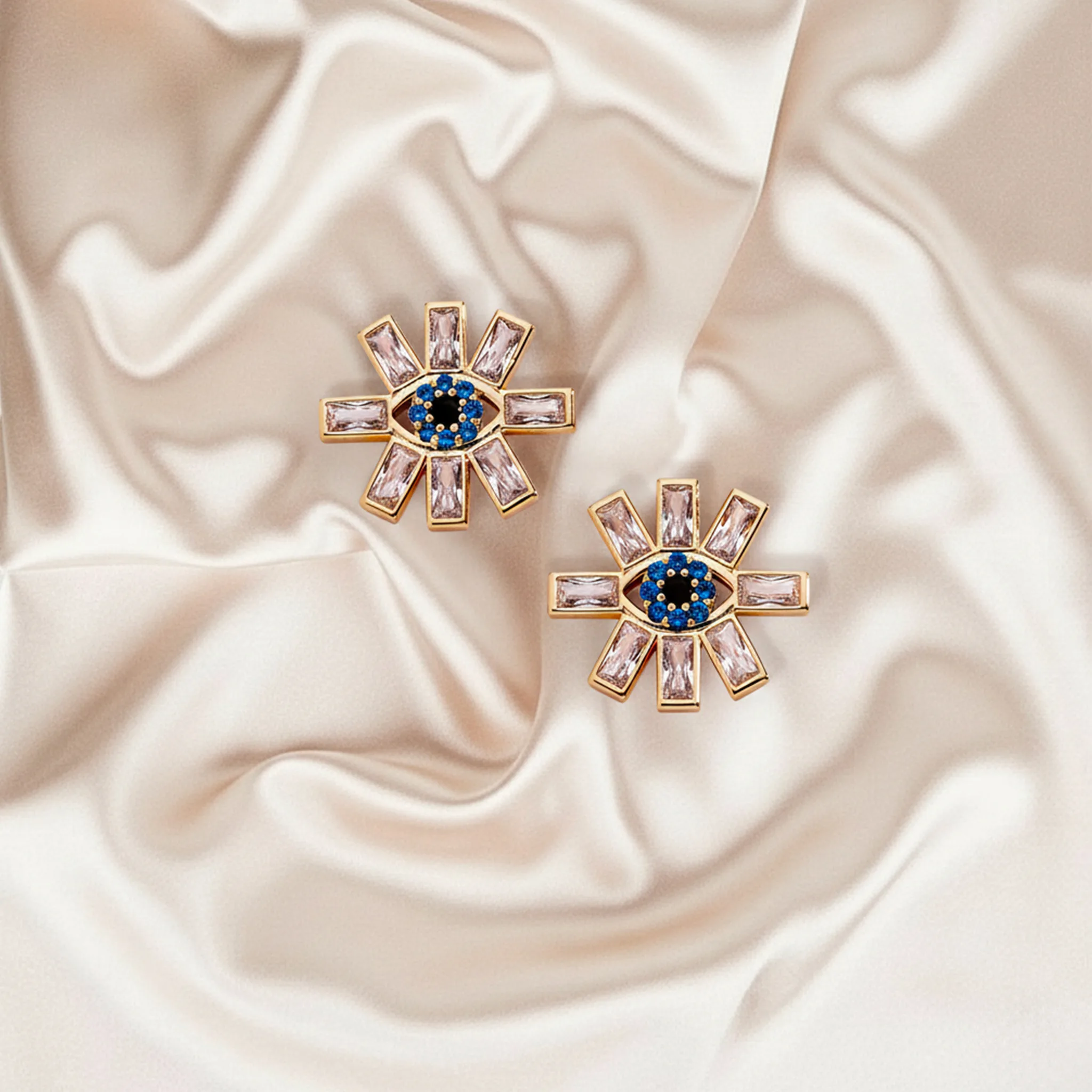 AI image of gold starburst earrings with pink baguette stones and blue evil eye centers on ivory satin. Alex and Ani AI Concept.