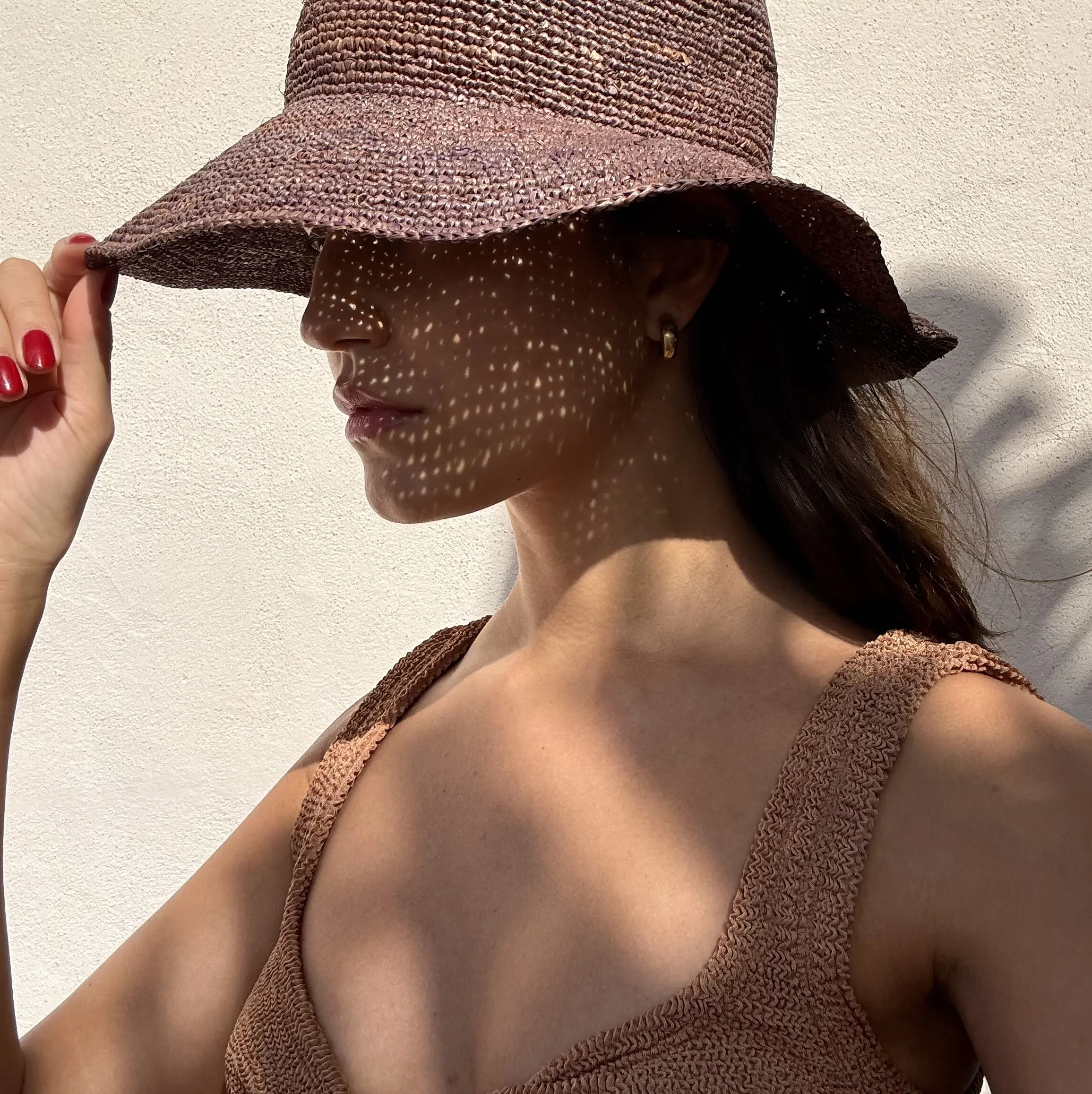 Woman in a brown crochet raffia sun hat and textured swimsuit, dappled sunlight across her face against a white wall.