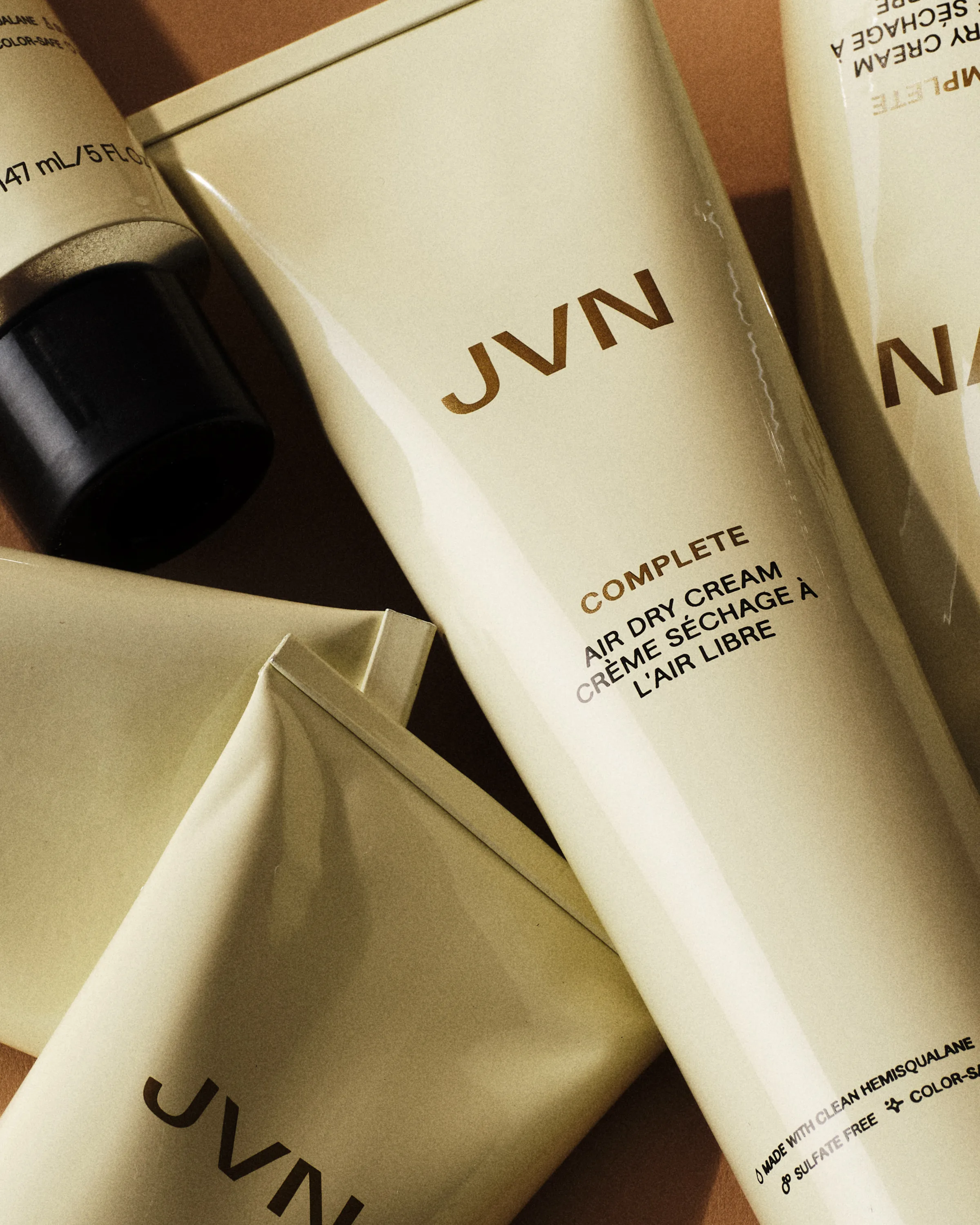 Cream-coloured Complete Air Dry Cream tubes piled together on a tan background, lit with hard shadows, for JVN.