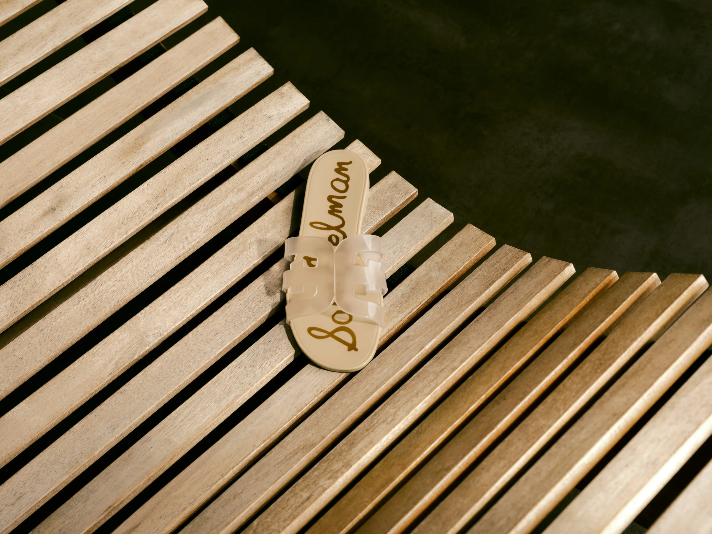 Clear jelly slide sandal with cut-out E straps resting on a curved wooden slatted bench, for Sam Edelman.