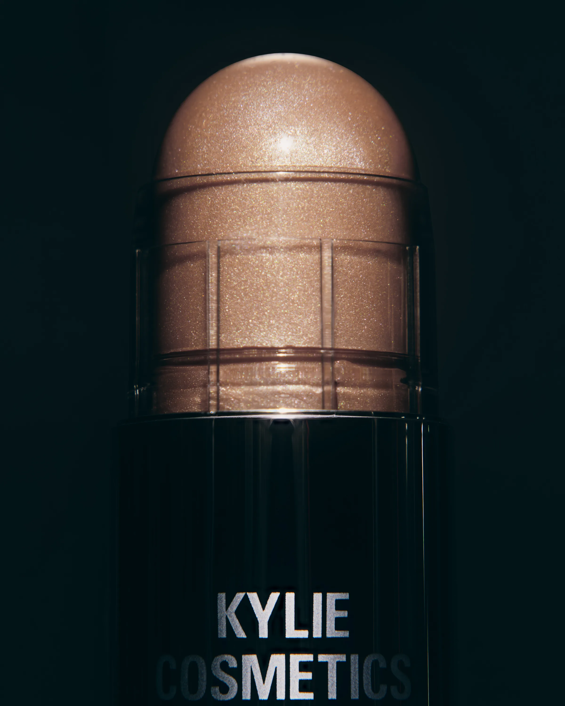 Close-up of a shimmering bronze Dewy Balm Stick in Lunar Sky on a black background, for Kylie Cosmetics.
