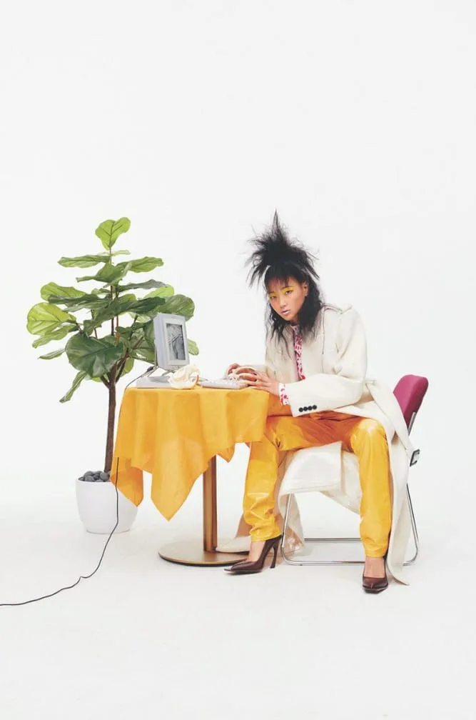 Model with spiked hair in cream coat and yellow leather pants types at a retro computer beside a fig plant, for Office Magazine.