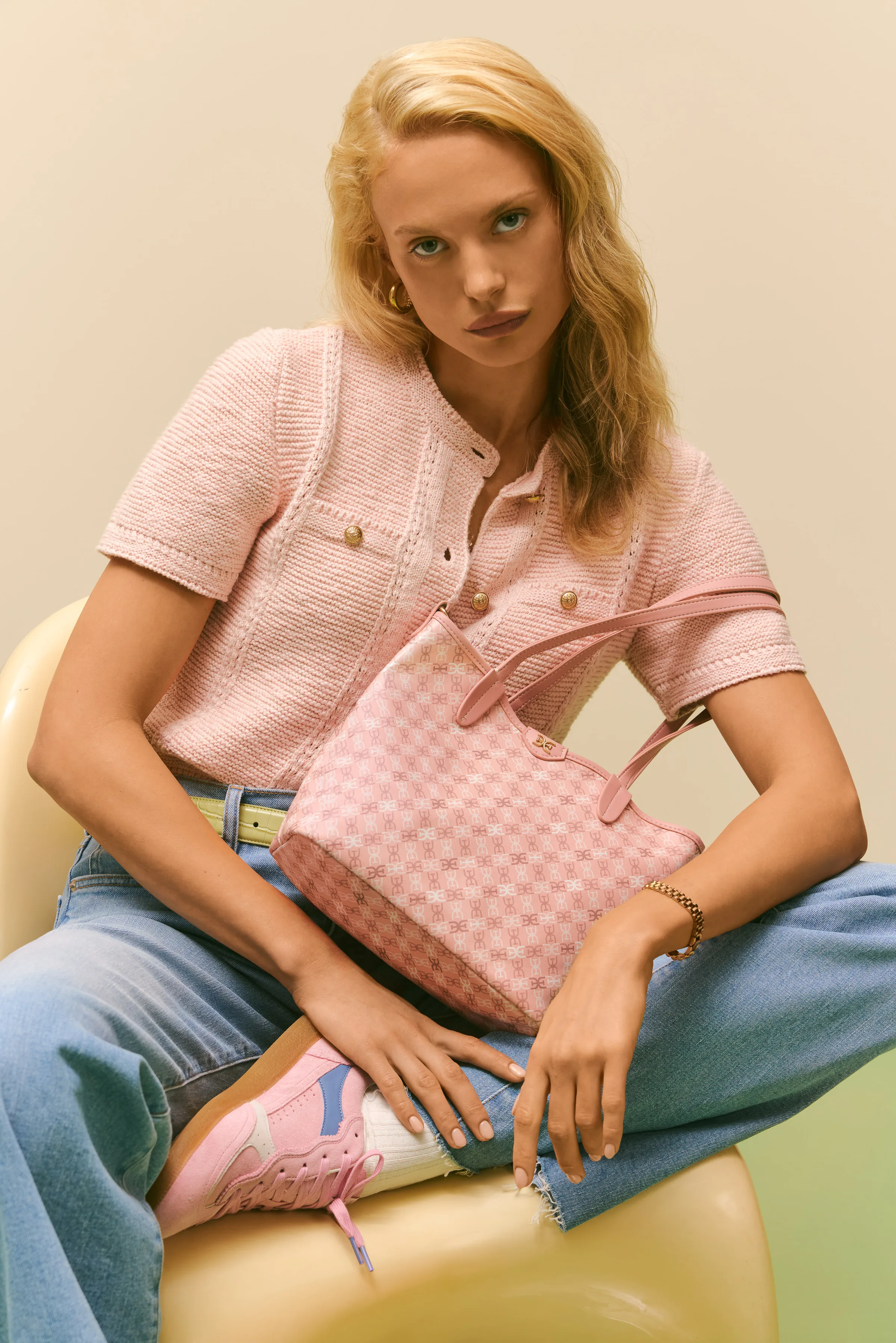Model in a pink knit top and jeans holding a pink monogram tote, wearing pink sneakers, seated on a chair, for Sam Edelman.