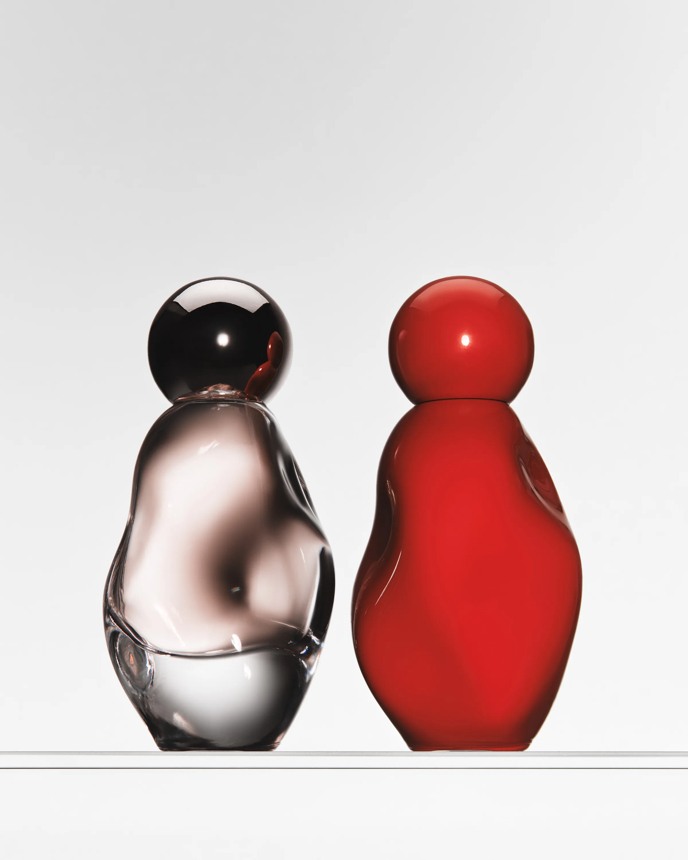 Clear and glossy red sculpted perfume bottles side by side on a glass shelf against a white backdrop, for Kylie Cosmetics.