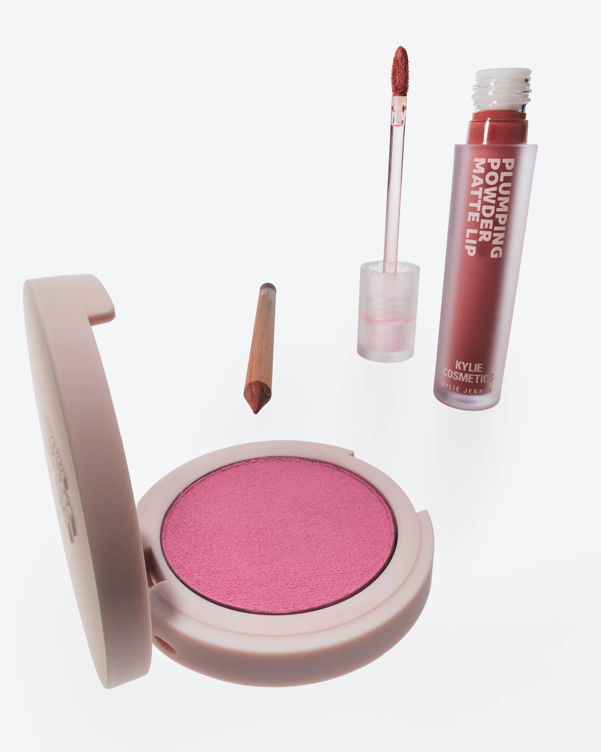 Open pink blush compact, lip liner pencil and plumping powder matte lip with wand floating on light grey, for Kylie Cosmetics.